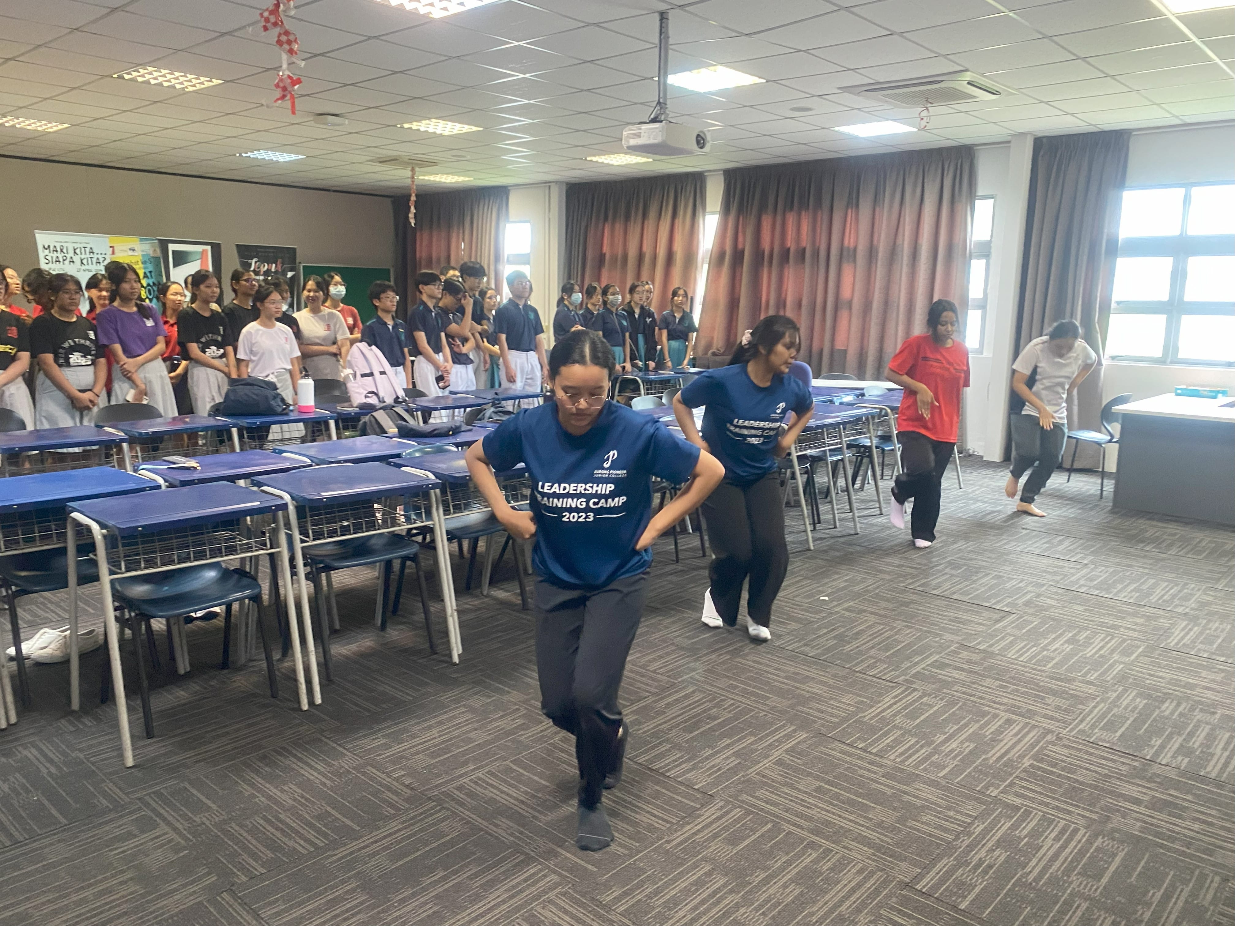 Students in blue Leadership Training Camp 2023 shirts perform a dance in a classroom while others watch.
