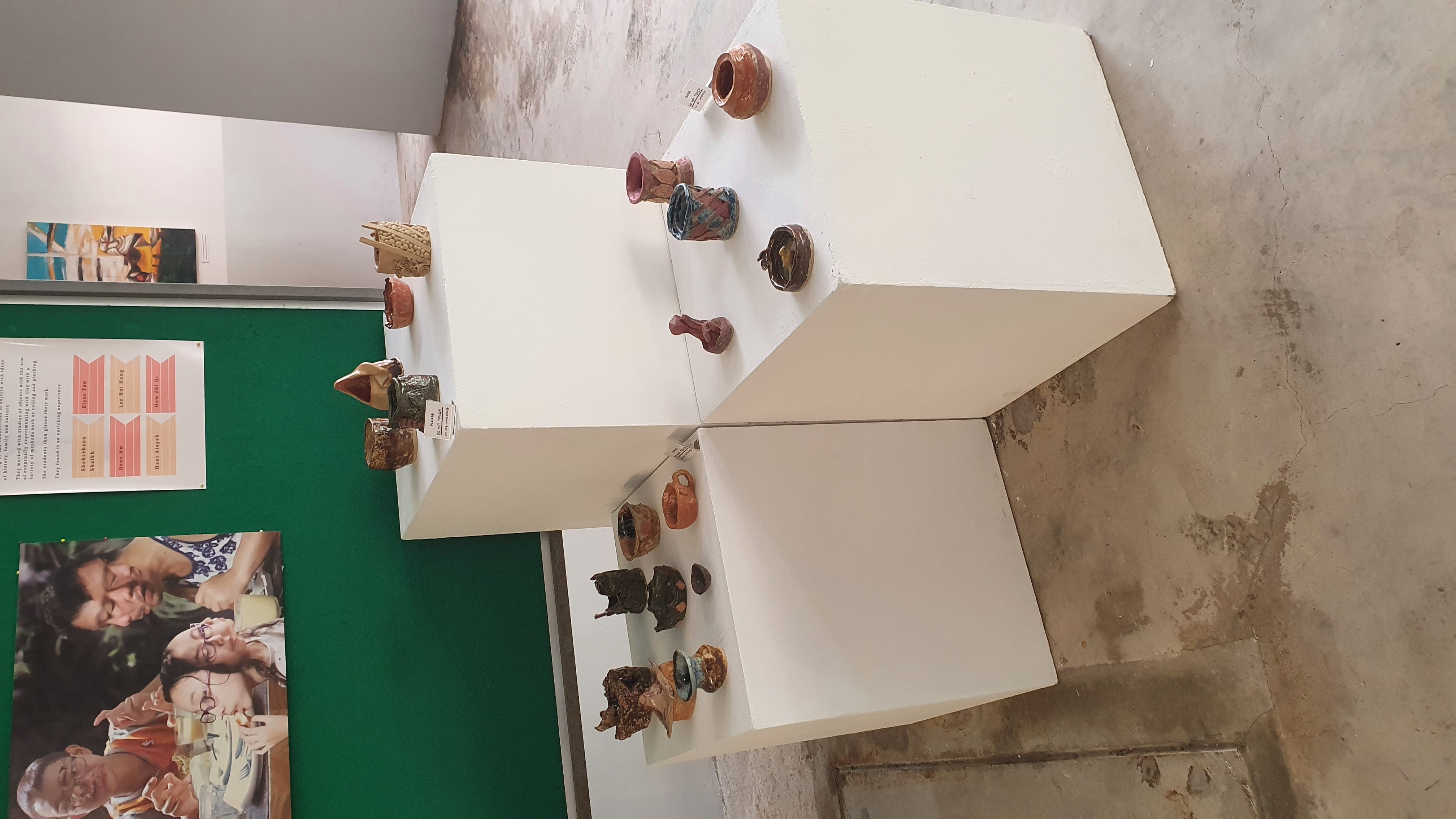 Three white pedestals displaying various small handmade ceramic bowls, cups, and sculptures in an art exhibition space.