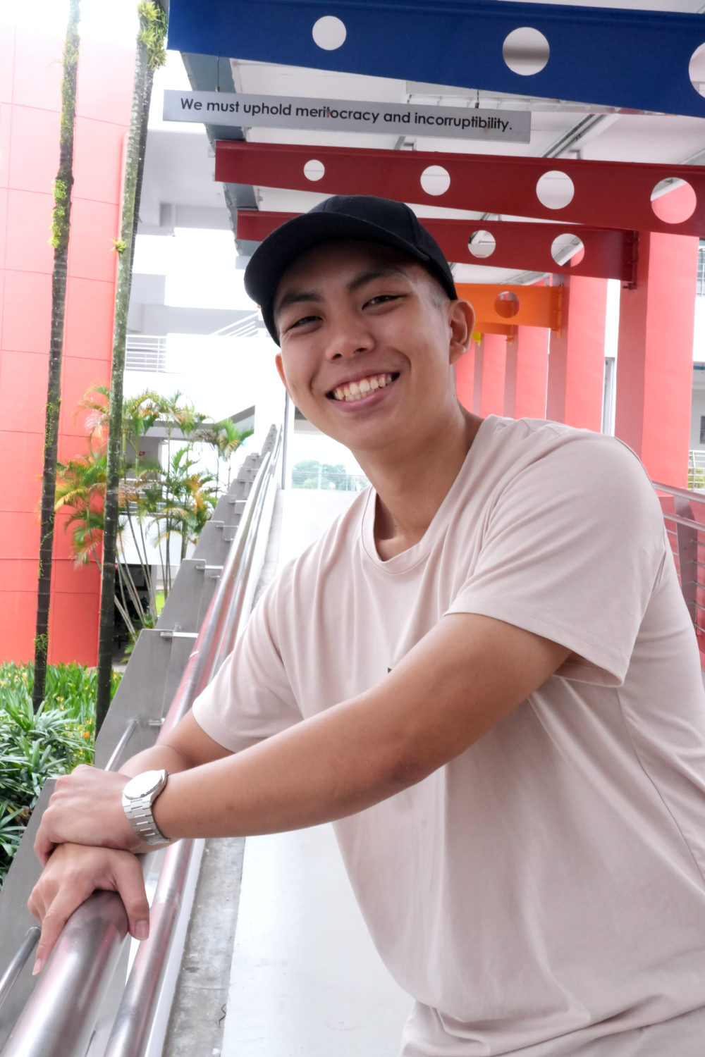 Young man in a black cap and pink t-shirt smiling, leaning on a railing outdoors near red and blue structures.