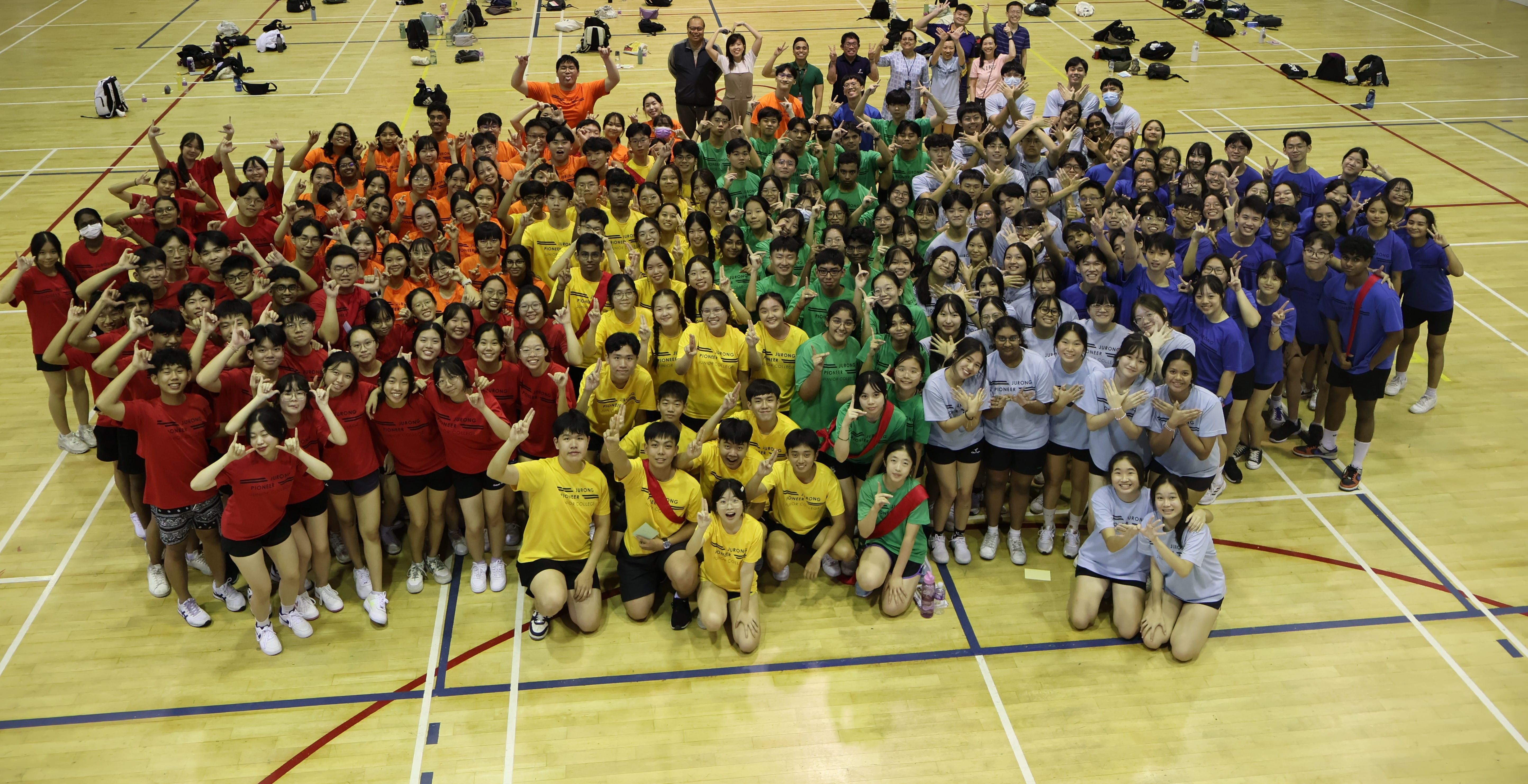 Large group of students in colored team shirts posing together on an indoor sports court.