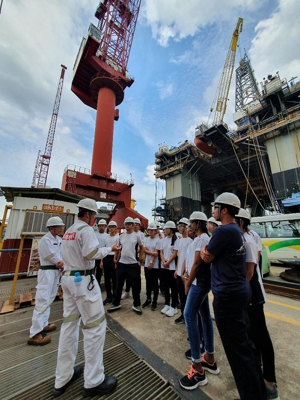 A group wearing white hard hats listens to a worker in white coveralls at an industrial shipyard with cranes.