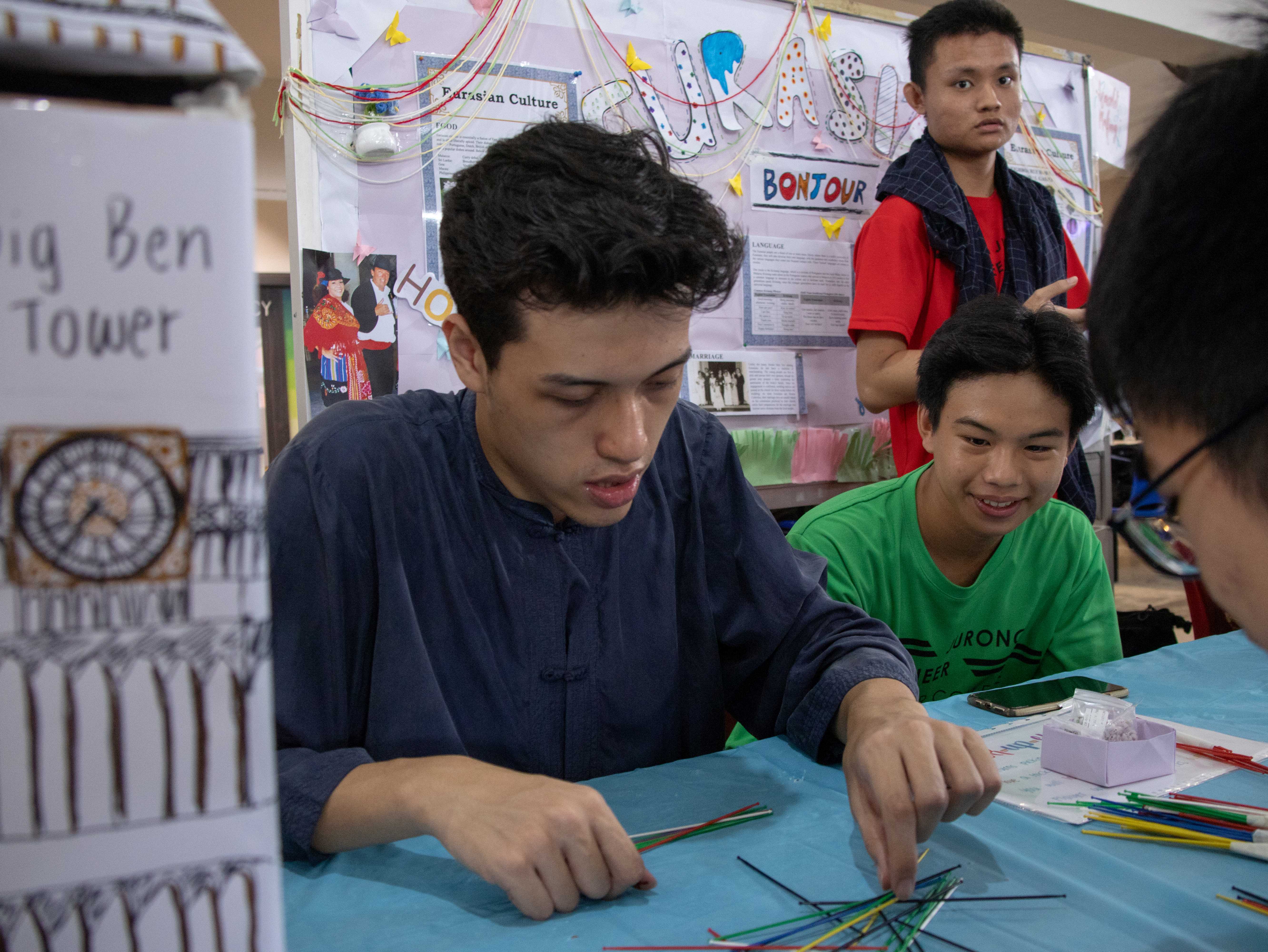 Three teenage boys at a table with colorful sticks at a school cultural fair with Eurasian Culture posters behind them.