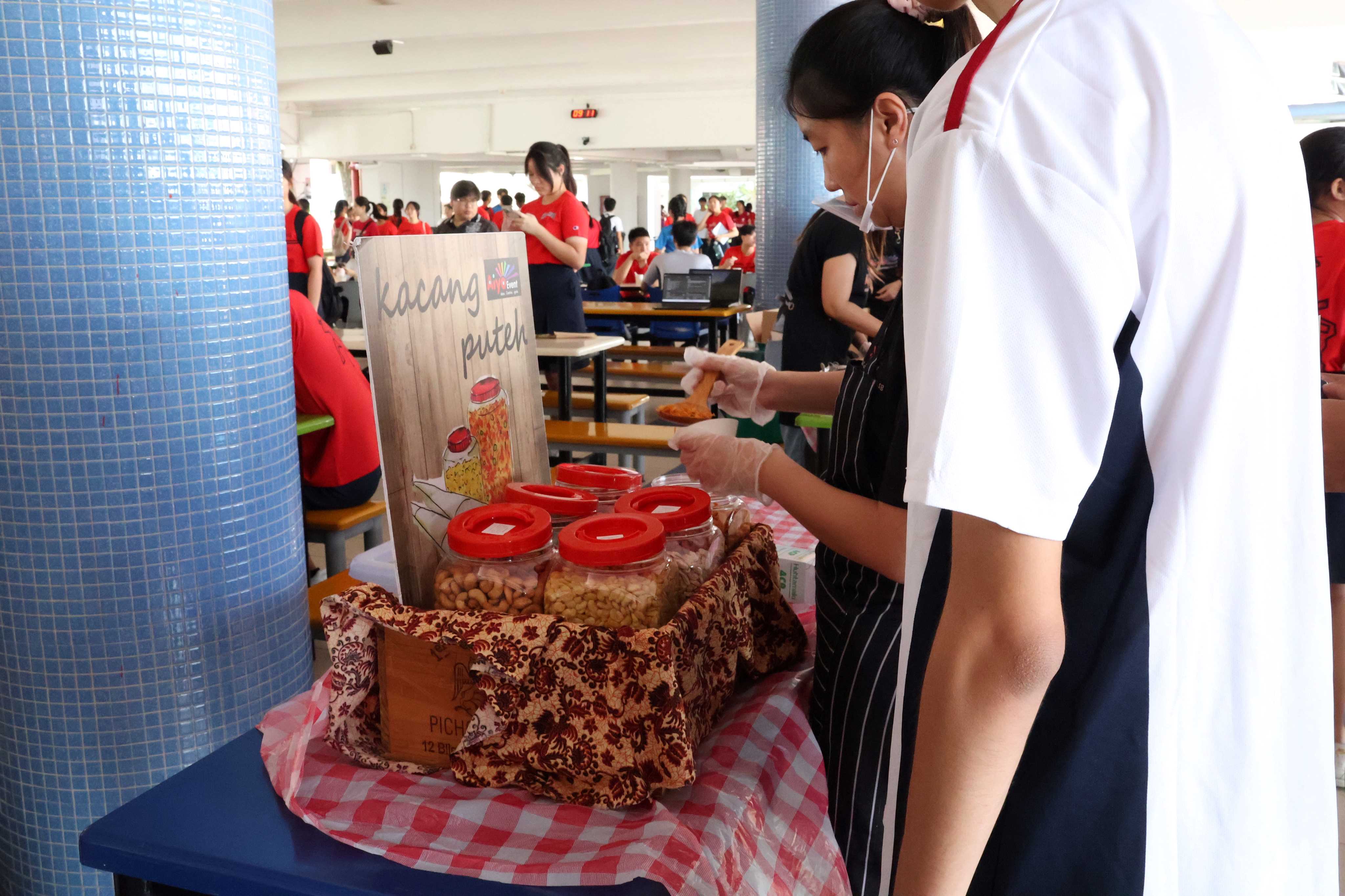 A student in an apron serves kacang puteh from jars with red lids at a school canteen stall.