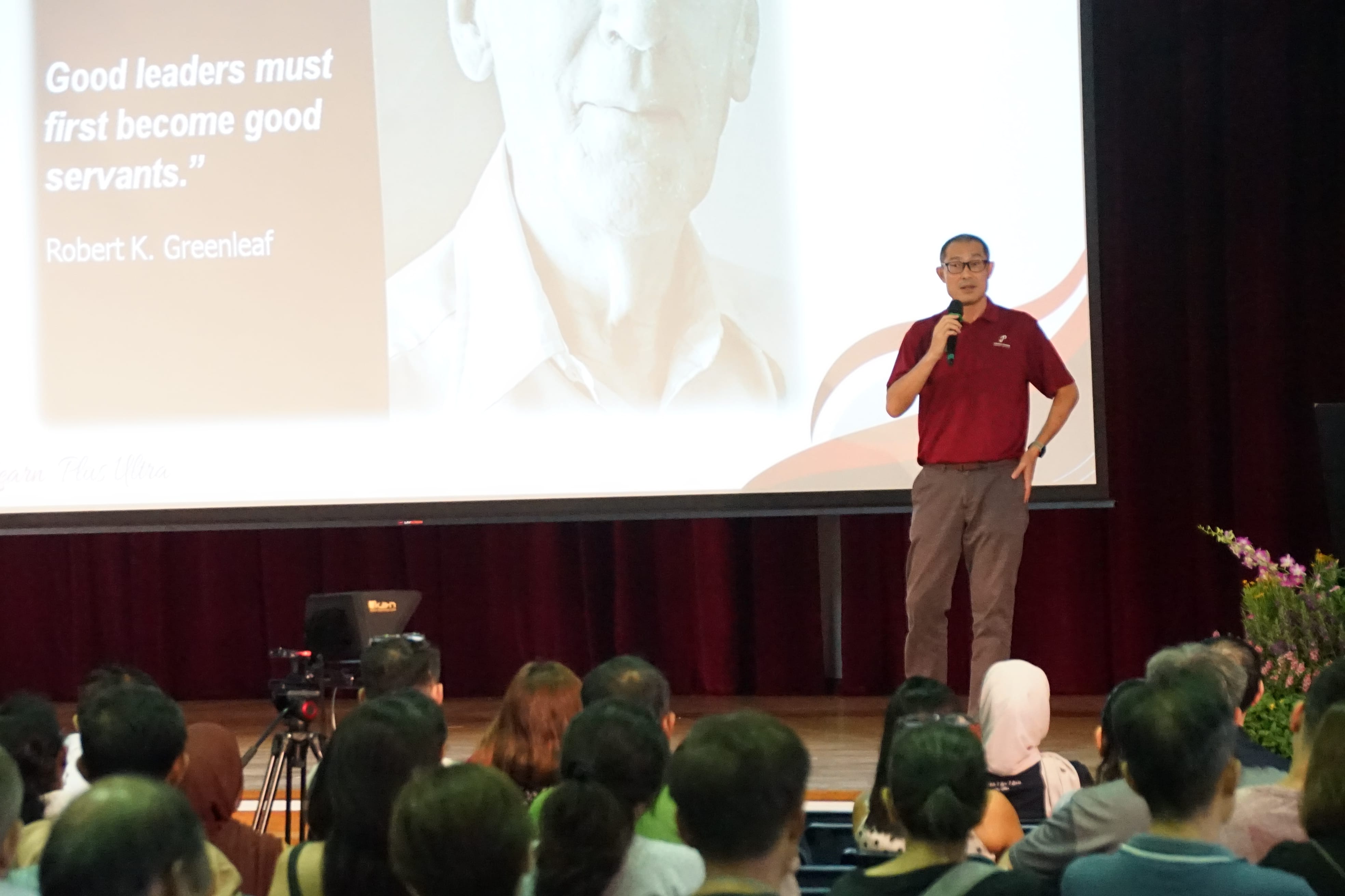 A man in a red shirt speaks into a microphone on stage before an audience, with a Robert K. Greenleaf leadership quote on screen.