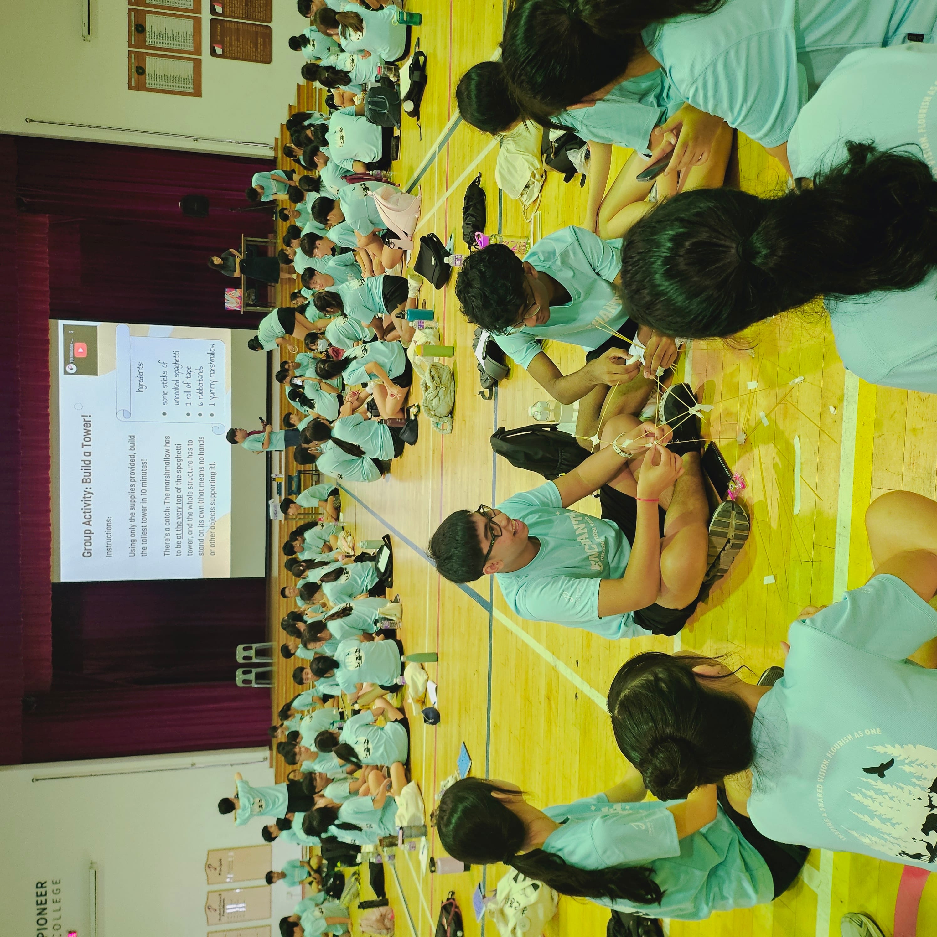 Students in matching teal shirts sit on a gym floor building spaghetti towers during a group activity session.