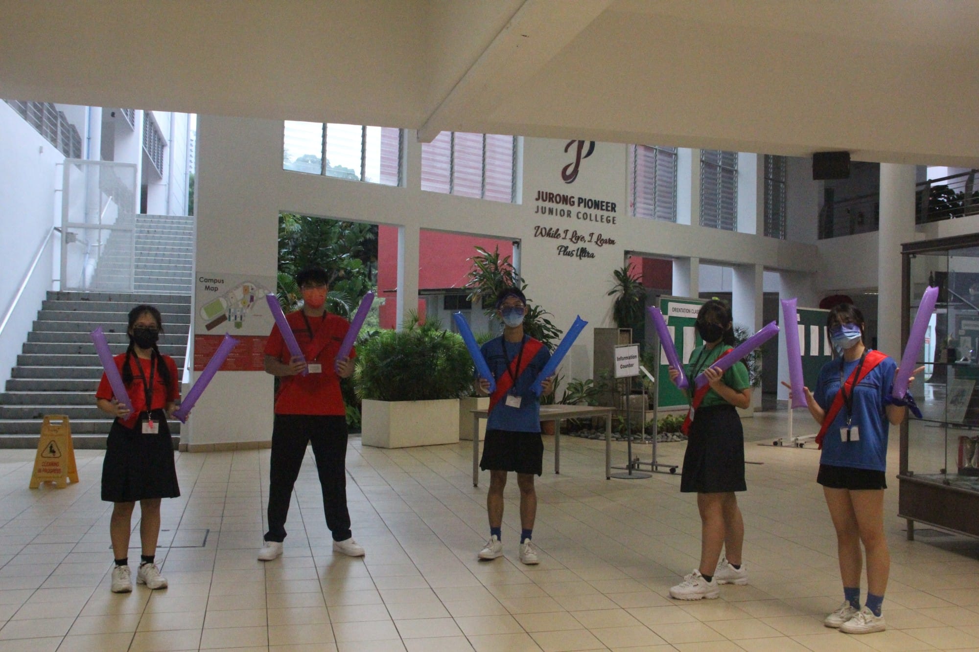 Five masked students hold colorful inflatable sticks in the atrium of Jurong Pioneer Junior College.