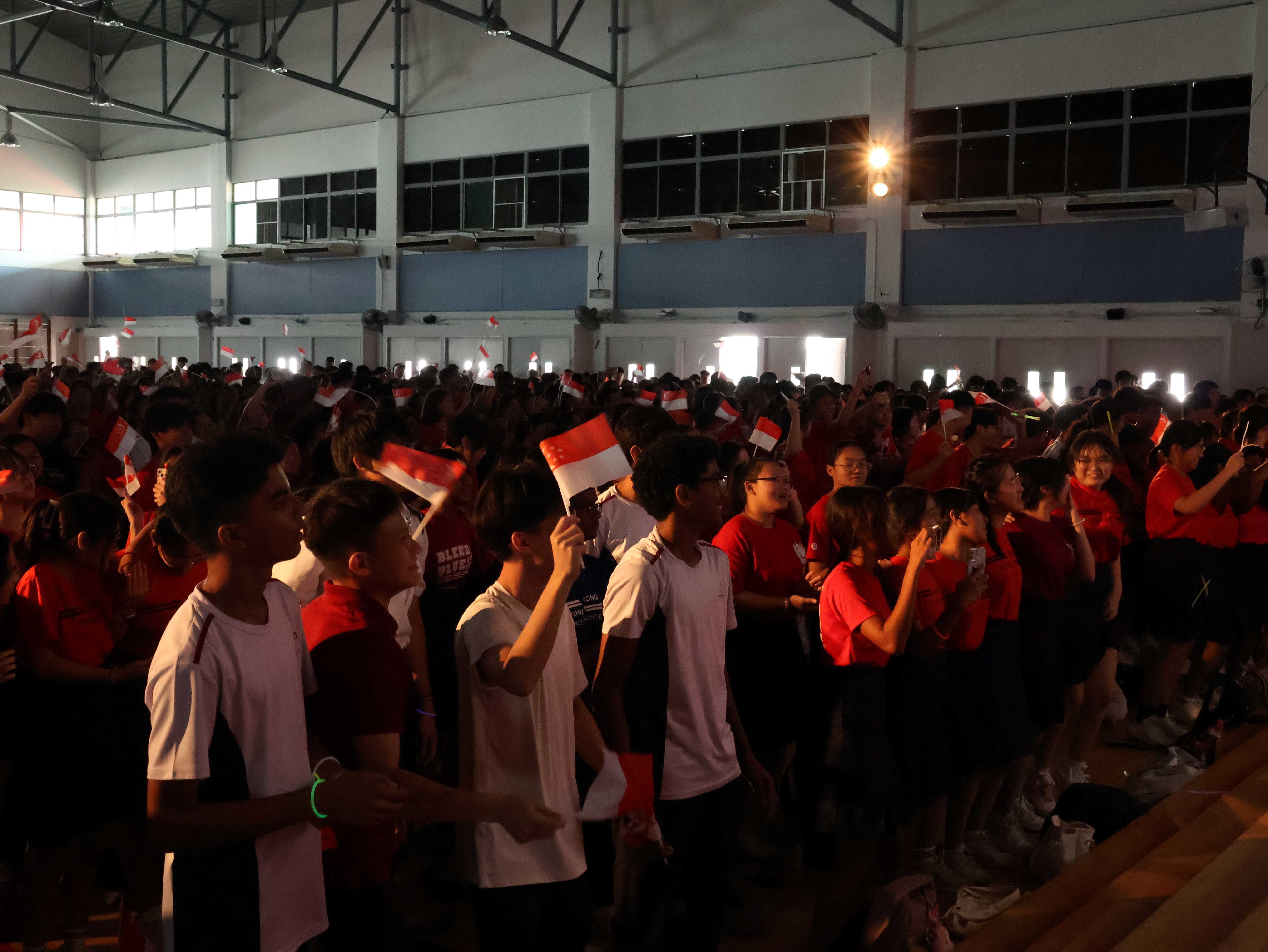 Large crowd of young people in red and white shirts waving Singapore flags inside a dimly lit hall.