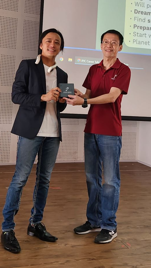 Two men smiling and holding a small box together in front of a projected JPJC Career Talk presentation slide.