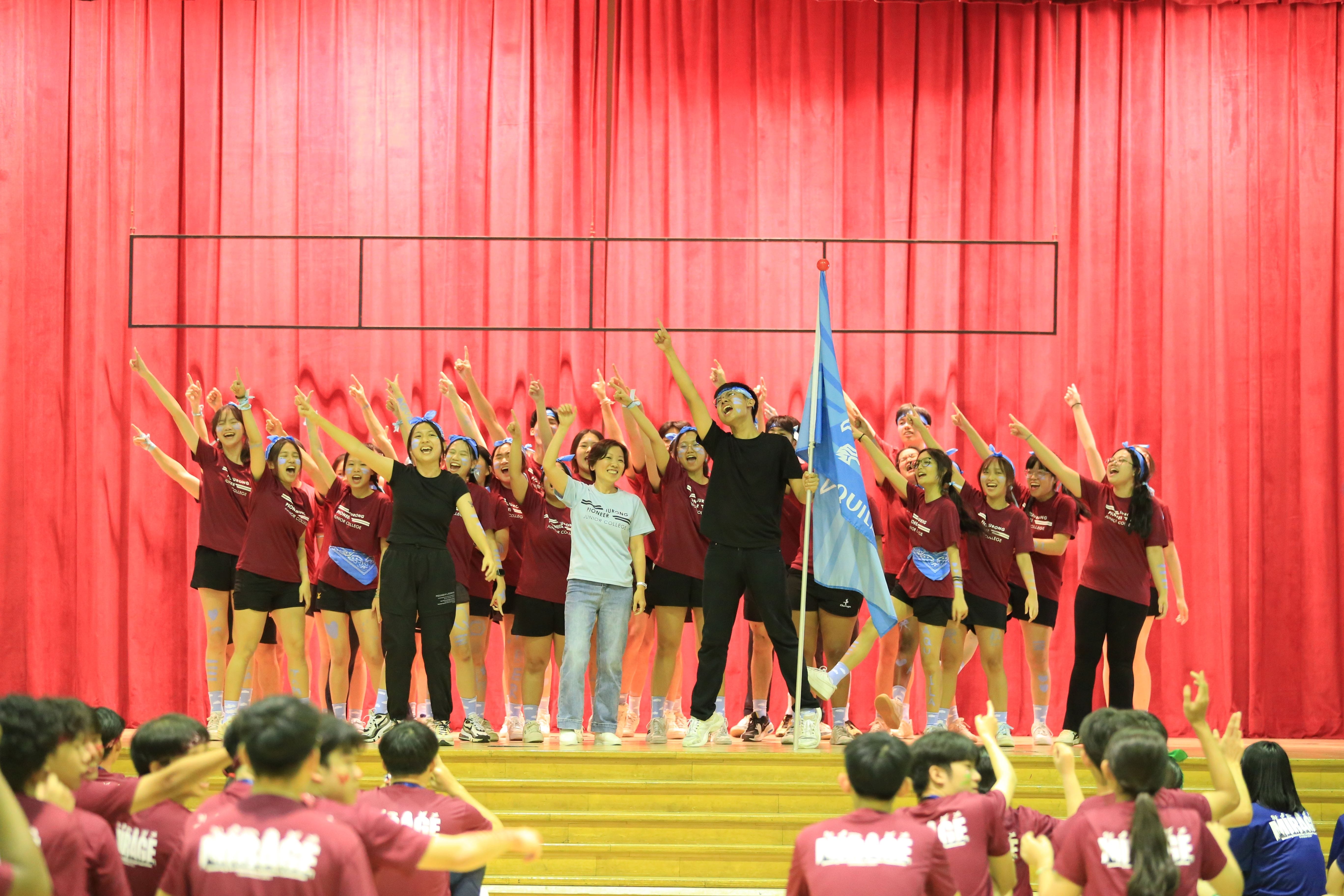 A large group of young people in maroon shirts cheer on a wooden stage before a red curtain, one holding a blue flag.