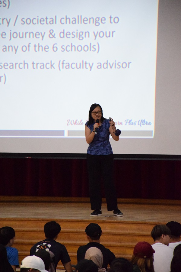 A woman in a blue top holds a microphone on stage, speaking to a seated audience with a presentation slide behind her.