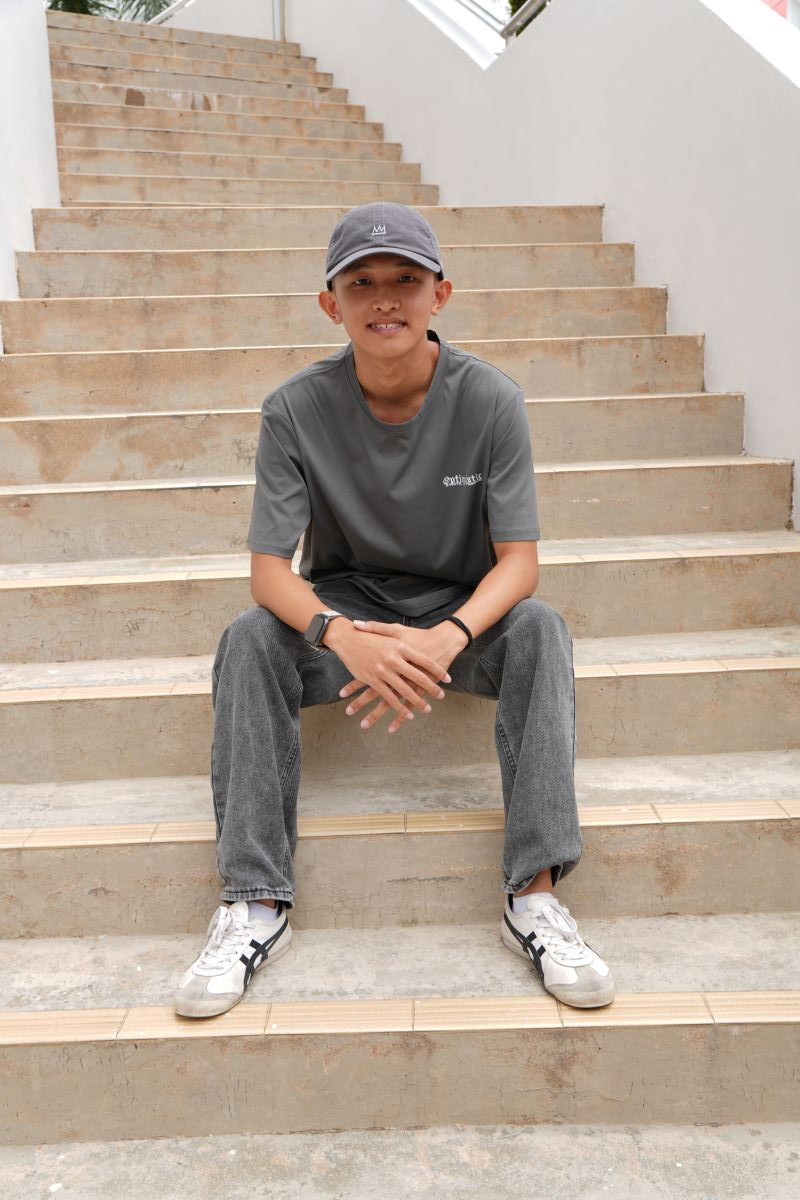 Young man in gray cap, t-shirt, jeans, and white sneakers sitting on outdoor concrete stairs, smiling at camera.