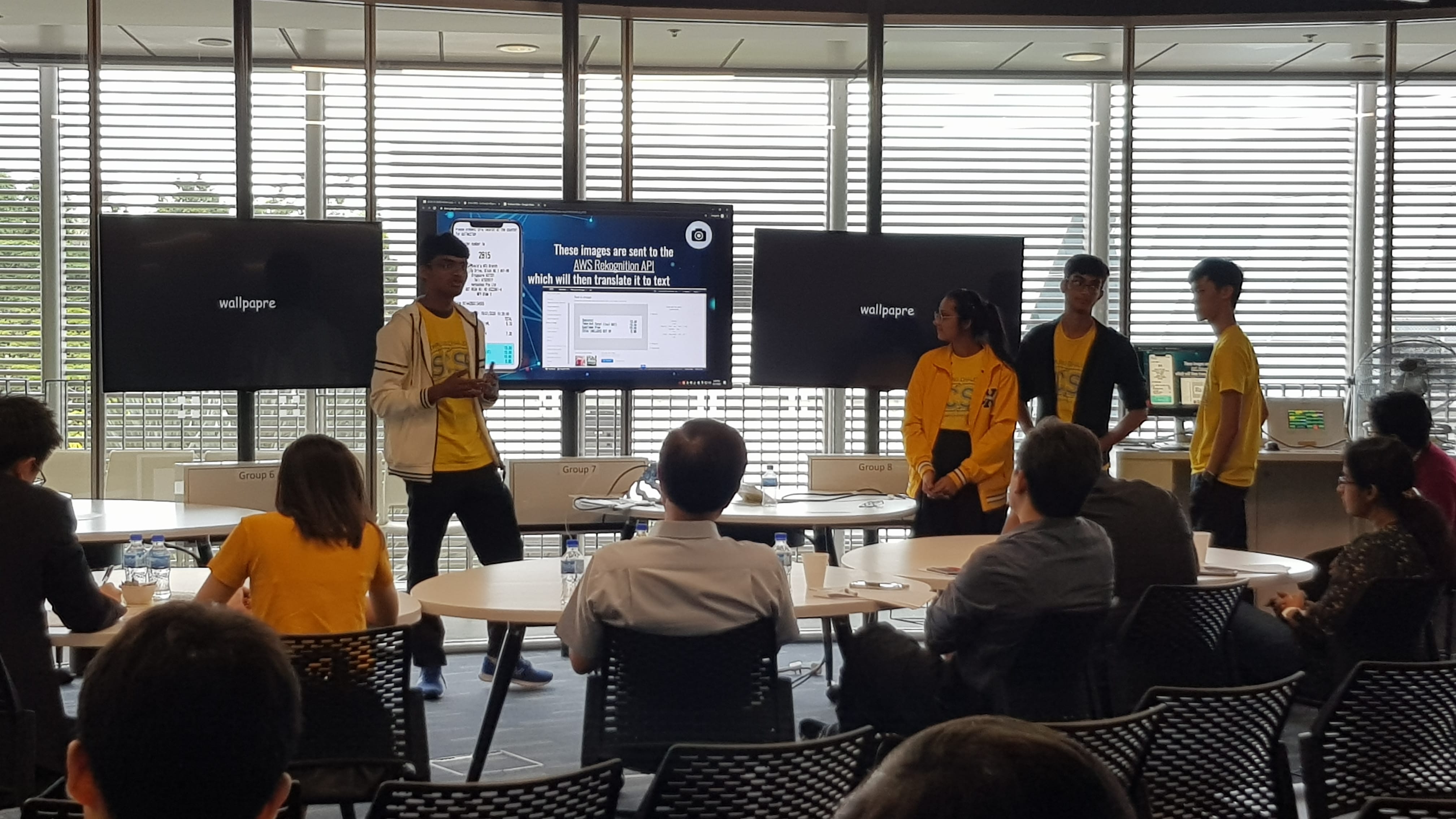 Students in yellow shirts present an AWS Rekognition API project on a large screen to a seated audience in a modern room.
