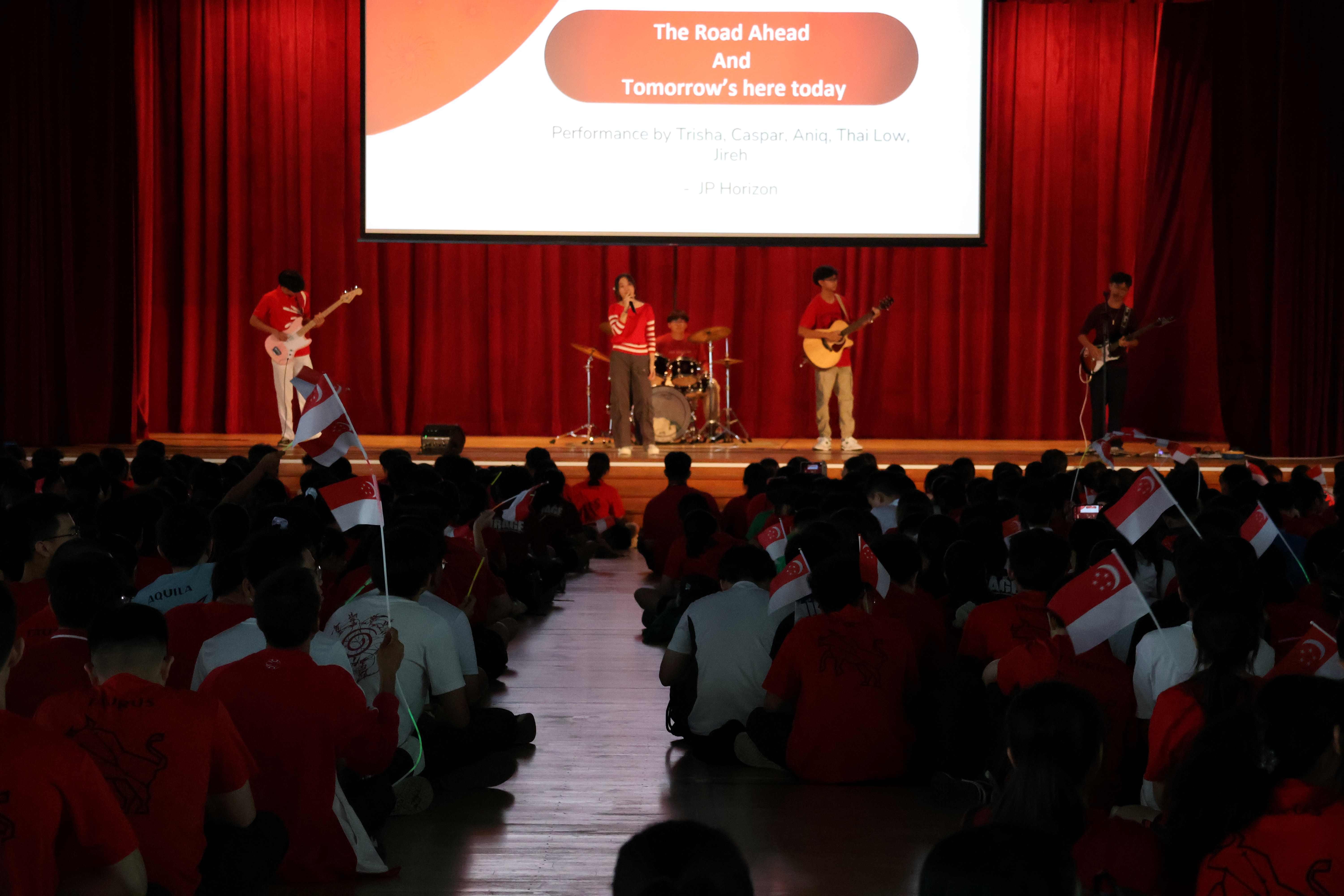 Students perform on stage with guitars and drums as a seated audience waves Singapore flags in a school hall.