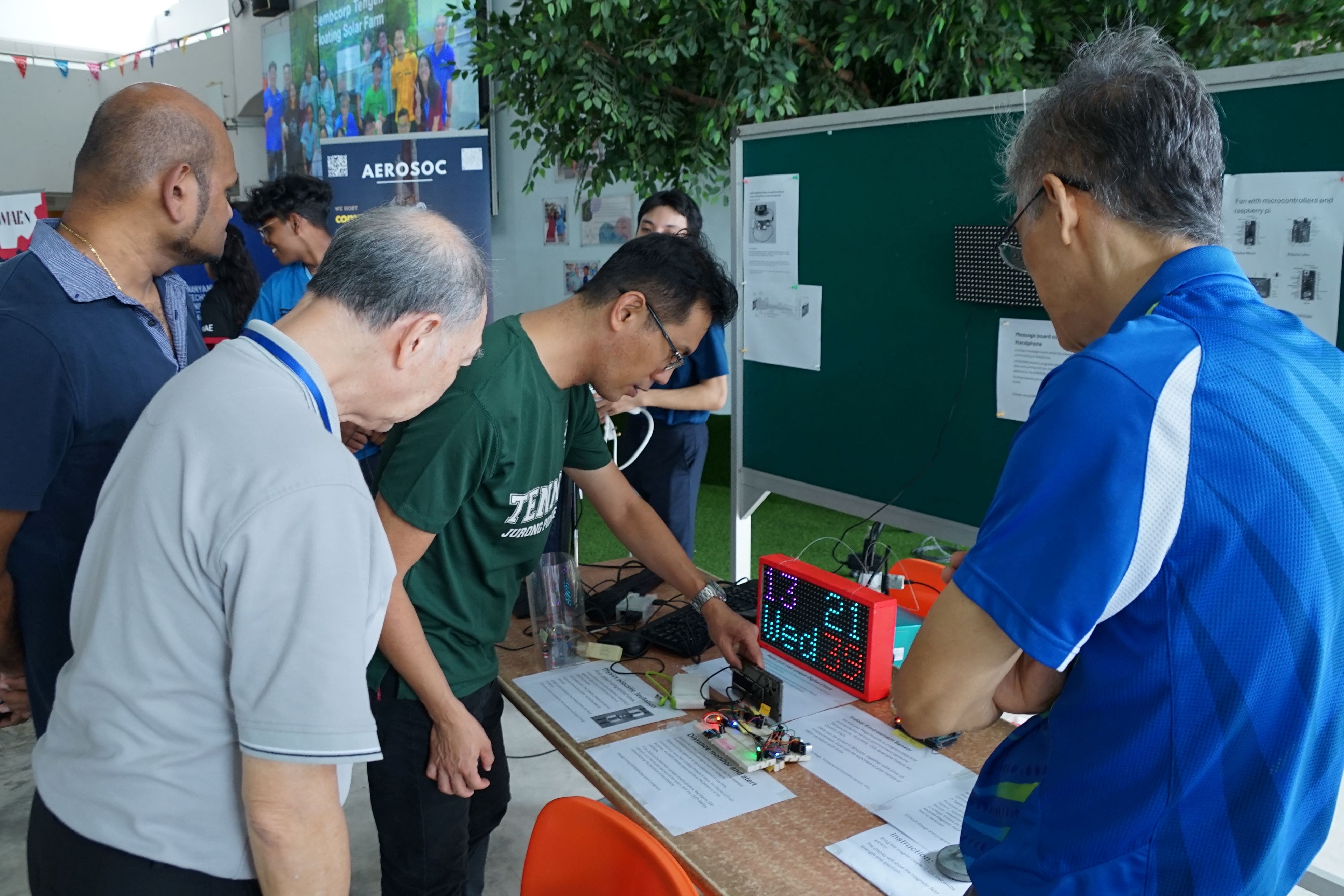 Several people gather around a table examining a red LED clock display and electronics at an indoor exhibition.