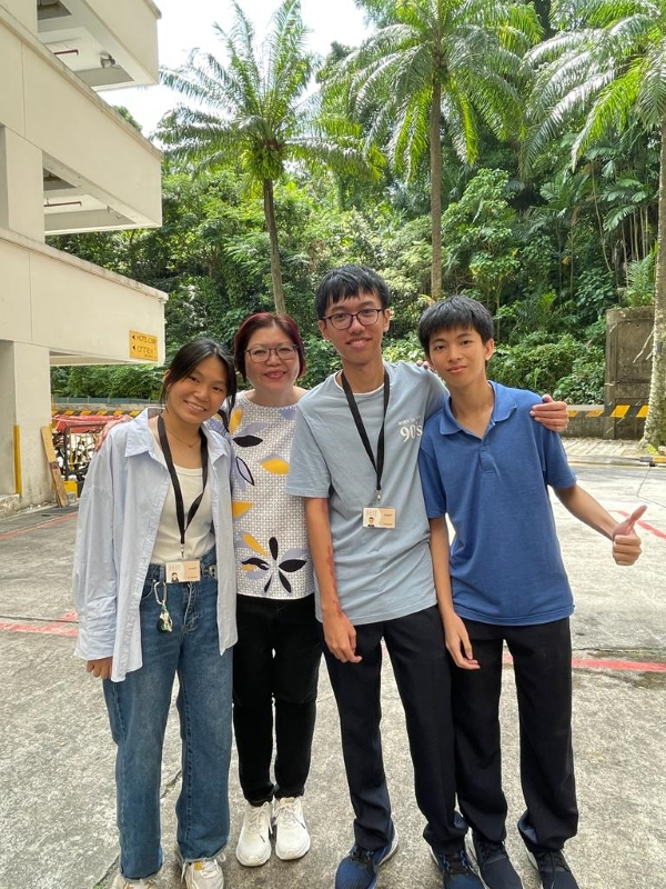 Four people smiling together outdoors near a building, with palm trees and green foliage in the background.