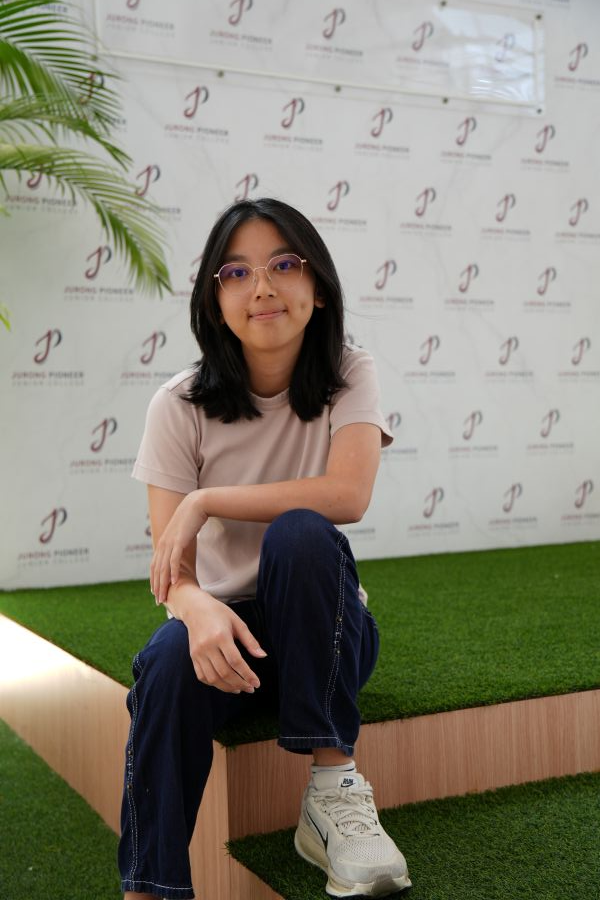 Young woman with glasses sitting on a grass-covered step before a Jurong Pioneer Junior College banner.