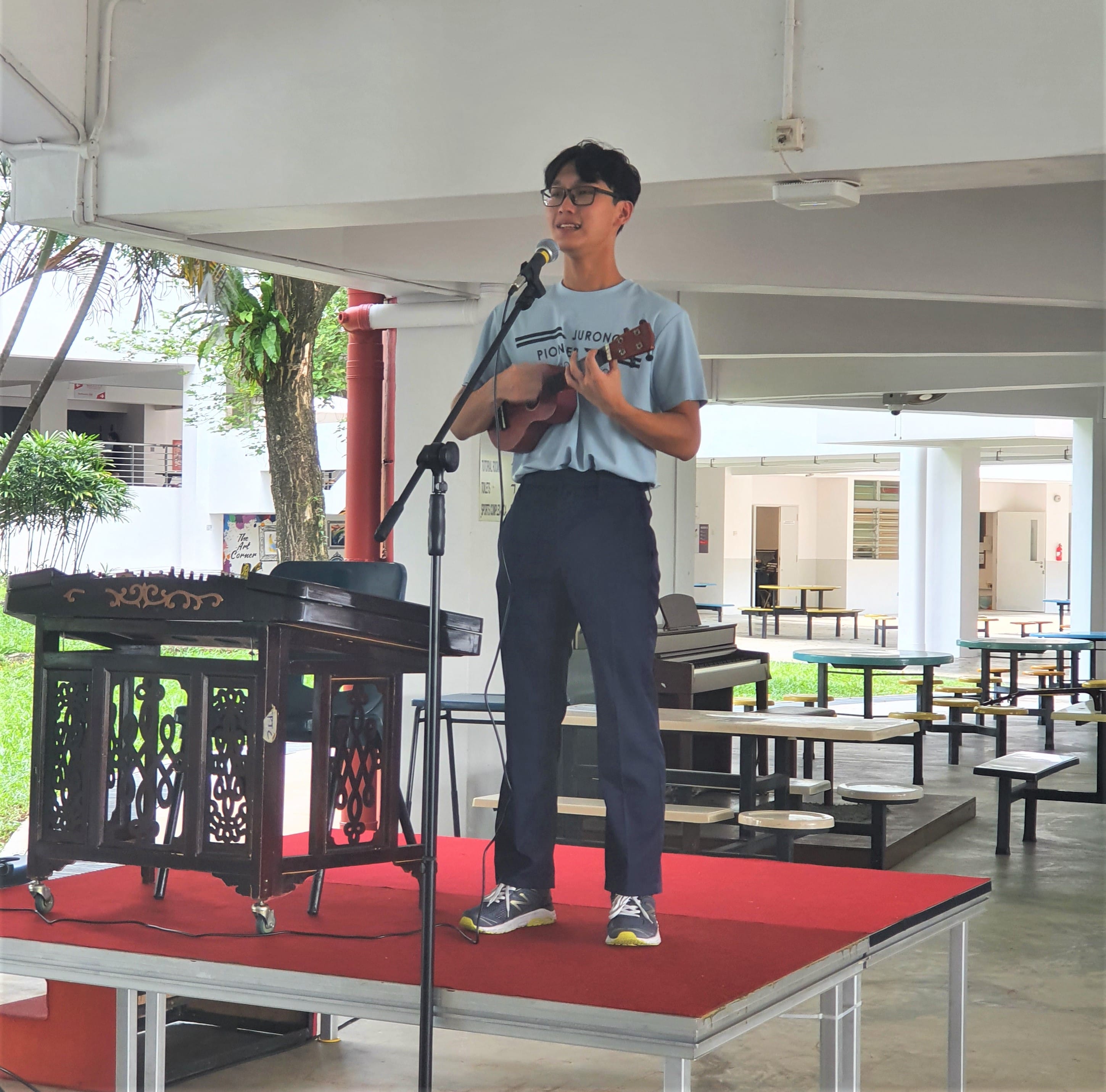 A young man wearing glasses plays a ukulele and sings into a microphone on a small red-carpeted stage.