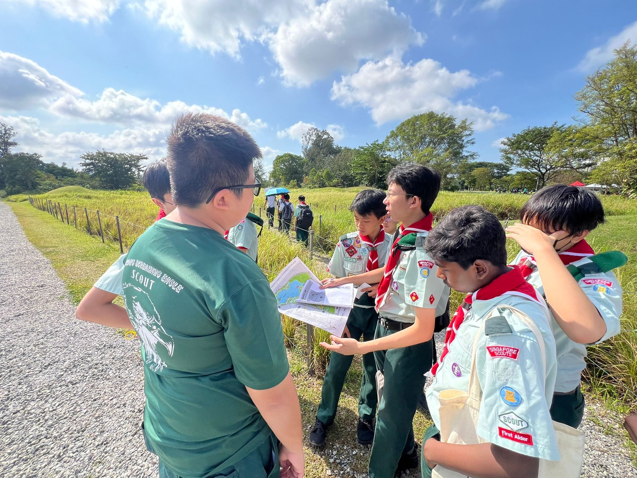 A group of Singapore Scouts in uniform huddle outdoors on a gravel path, reading a map on a sunny day.