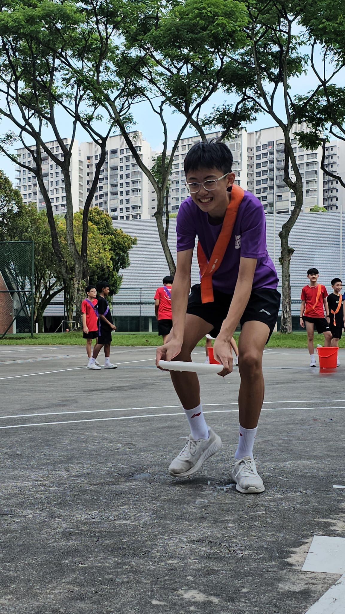 A smiling teen in a purple shirt holds a white tube during an outdoor game on a court with other students in the background.