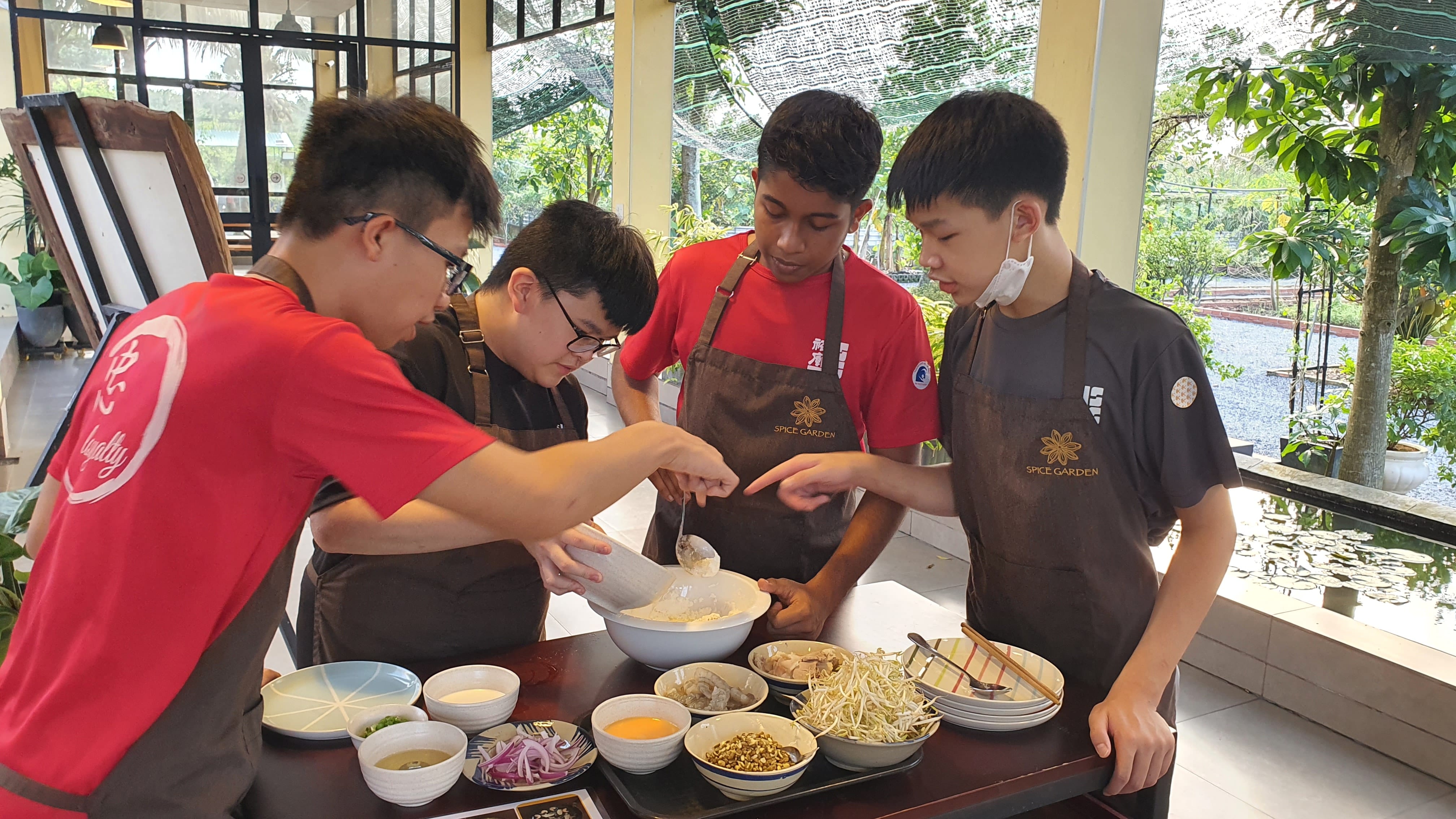 Four teens in aprons mix ingredients in a bowl at a table with small dishes in a bright room.
