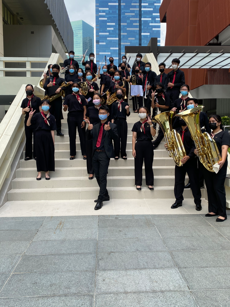 A large youth band in black uniforms and face masks poses on outdoor steps holding brass instruments.