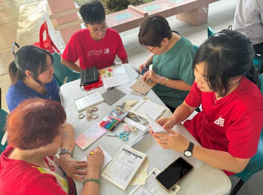 Five people sit around a table doing a craft activity with paper, scissors, tape, and markers outdoors.
