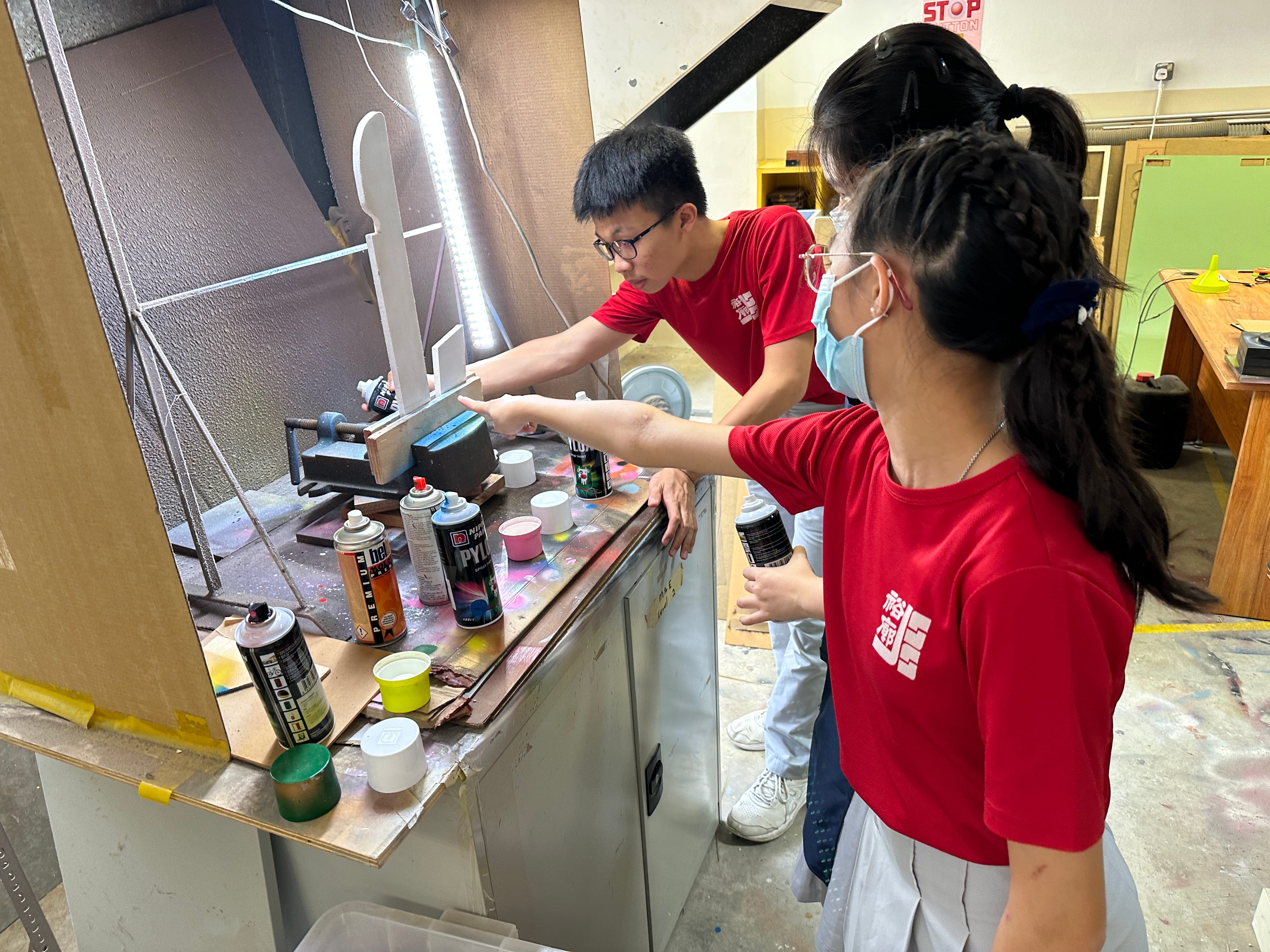 Two students in red shirts work at a paint-covered workbench with spray cans and a lit booth in a workshop.