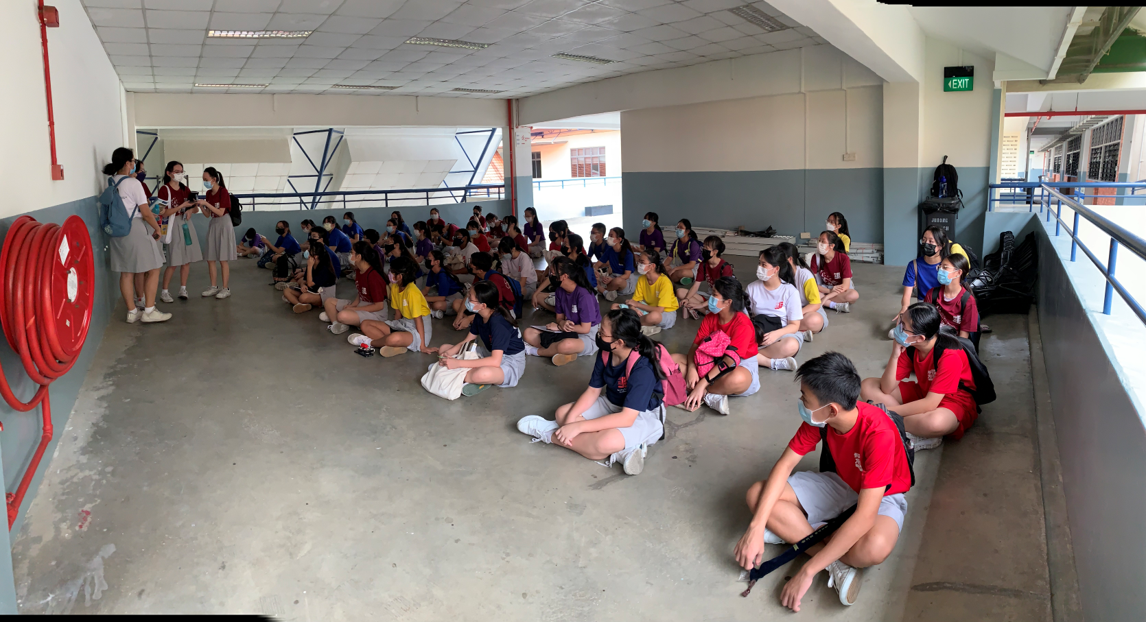 A large group of masked students in colorful shirts sit on a concrete floor in a covered school corridor.