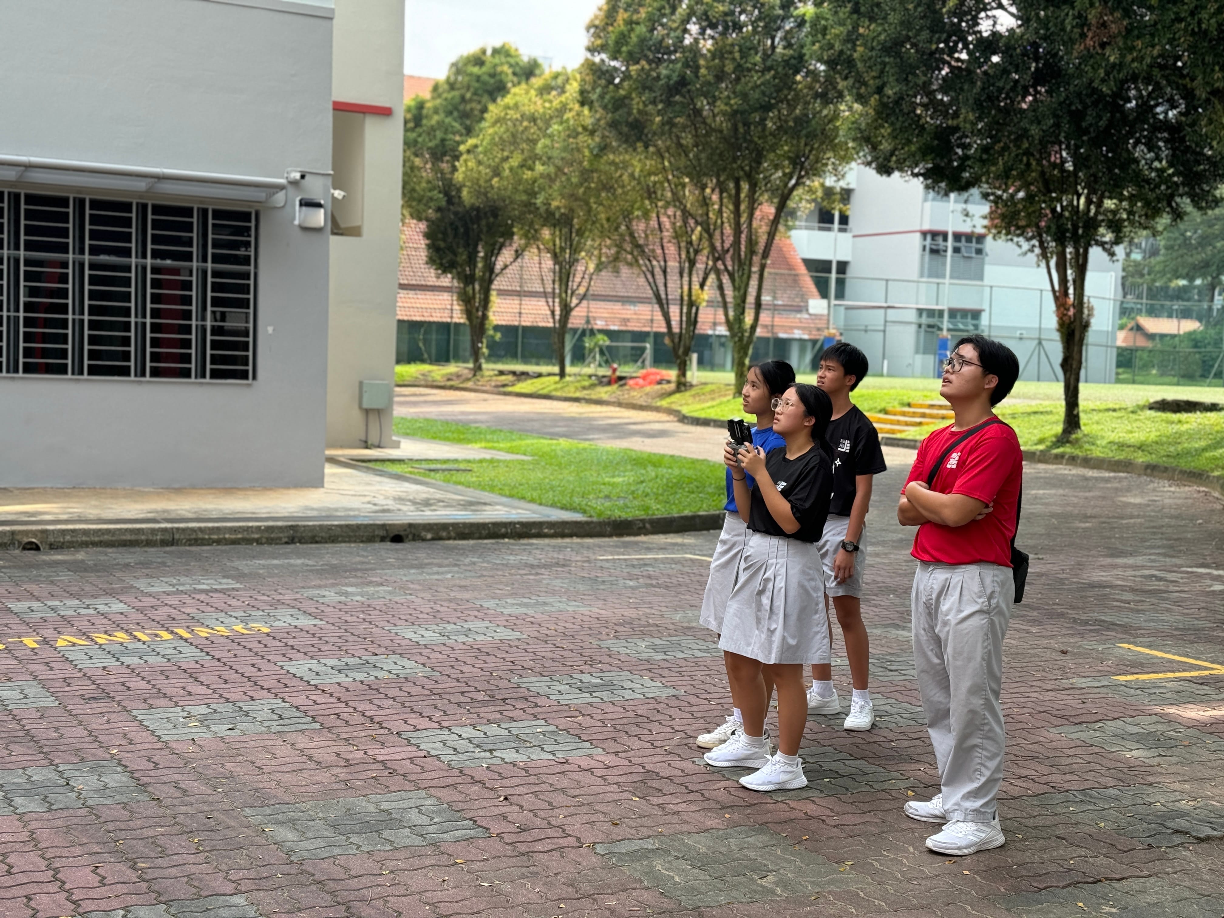 Four students in school uniforms stand on a brick courtyard, looking upward near a grey building surrounded by trees.