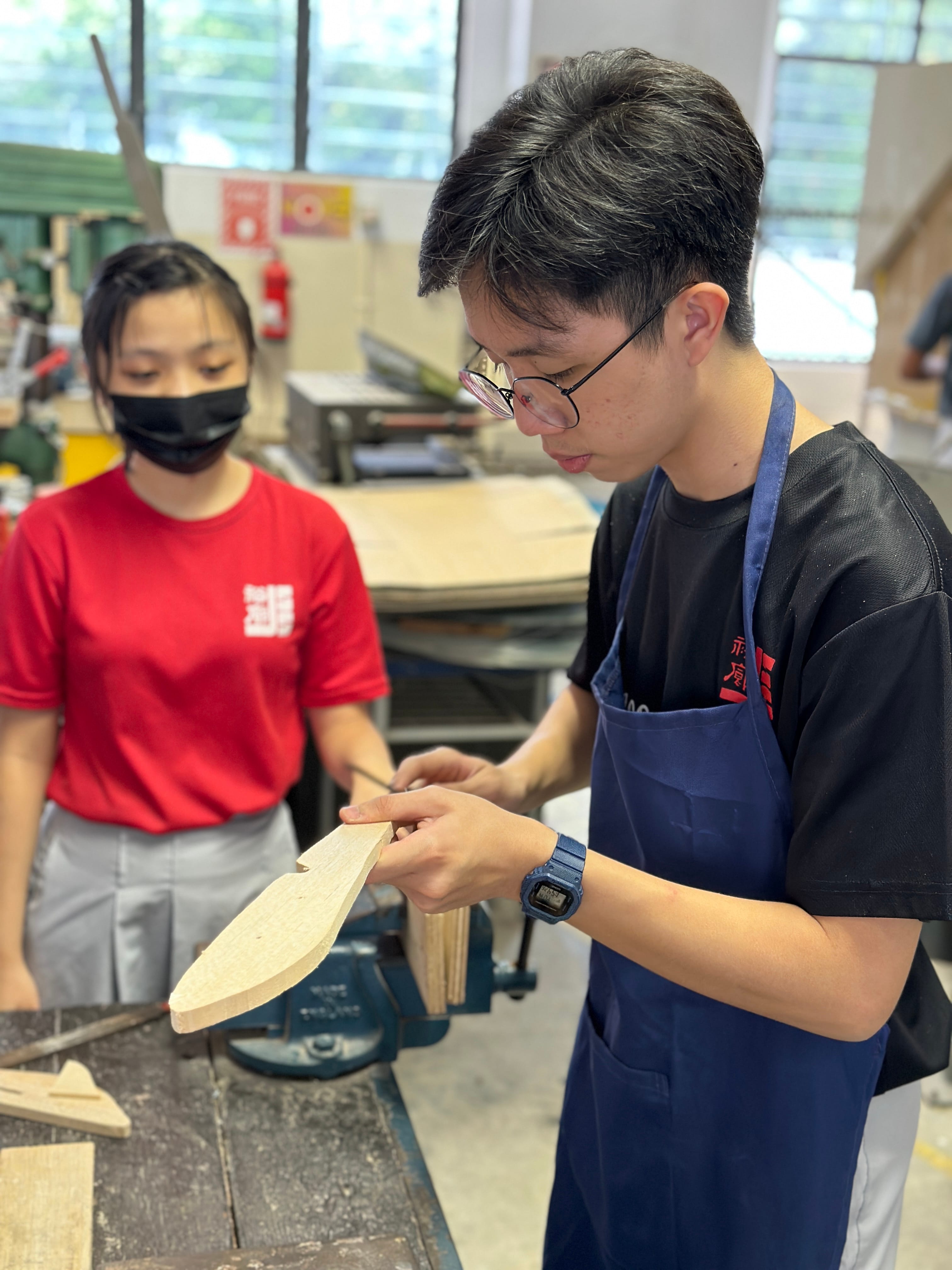 A young man in a blue apron examines a wooden piece in a workshop while a masked girl in red watches.