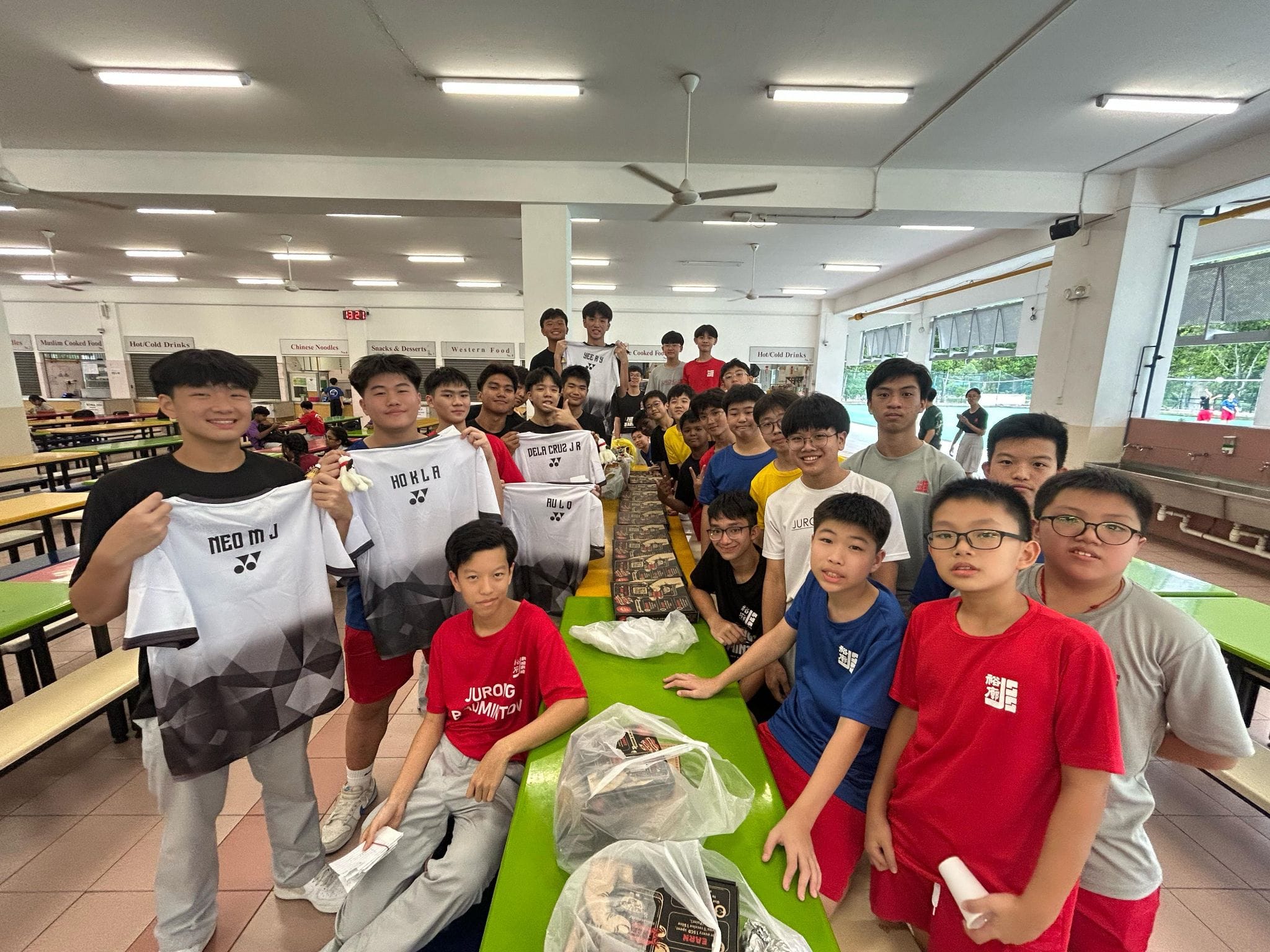 A group of teenage boys in a school canteen, some holding white Yonex badminton jerseys with name prints.