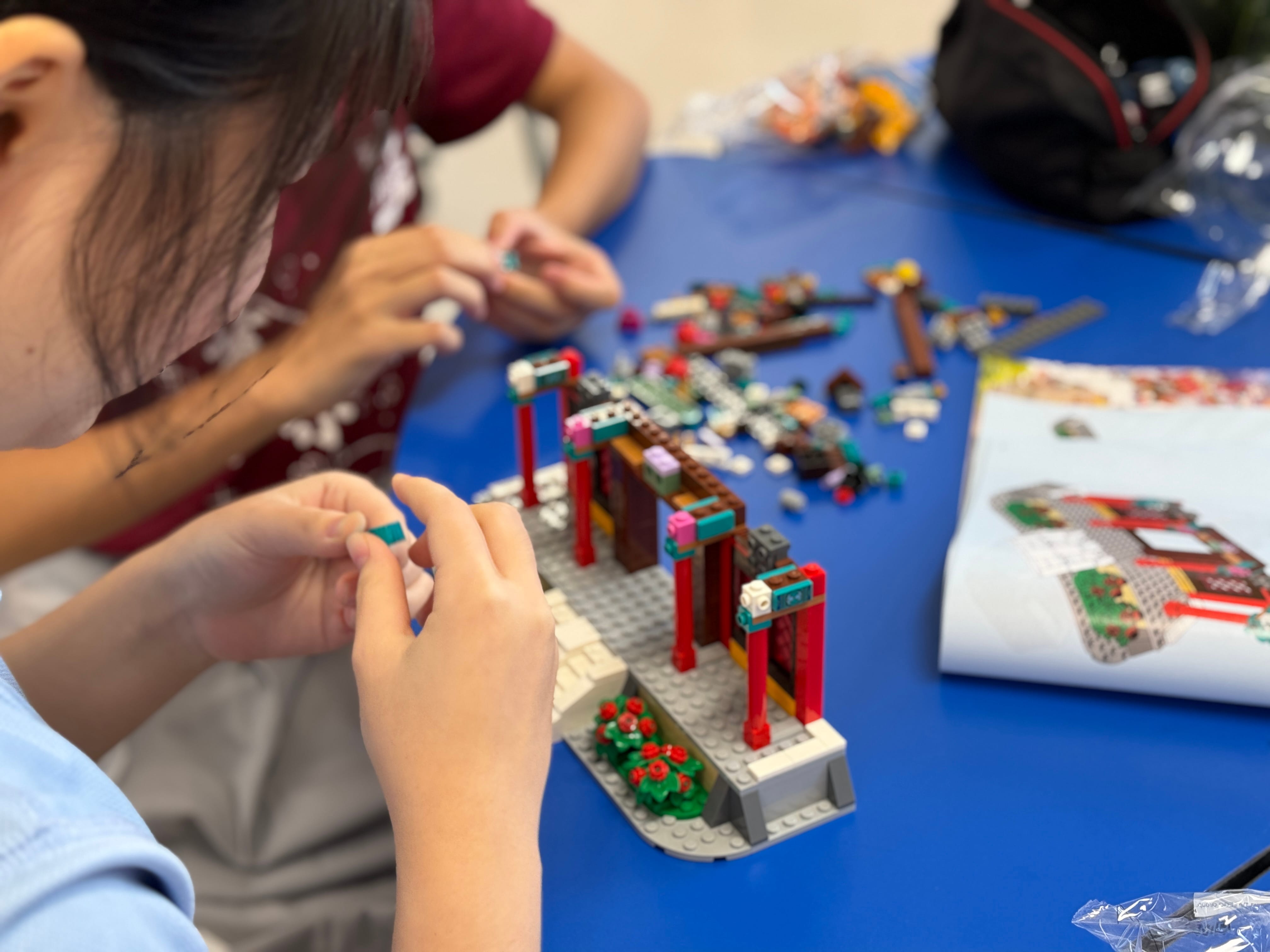 Two people build a colorful LEGO set with red pillars on a blue table, with loose bricks and instructions nearby.