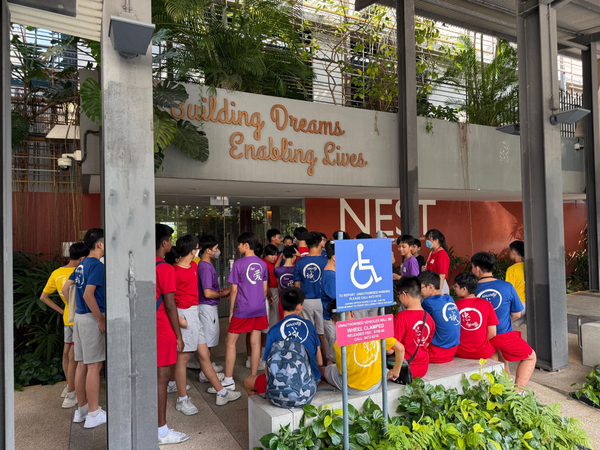 Students in colorful shirts gather outside a building entrance with a sign reading "Building Dreams Enabling Lives" and "NEST."