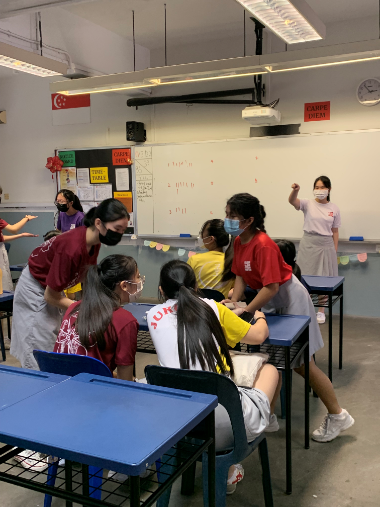 Students wearing masks gather around desks in a classroom with a whiteboard, colorful posters, and a Singapore flag.