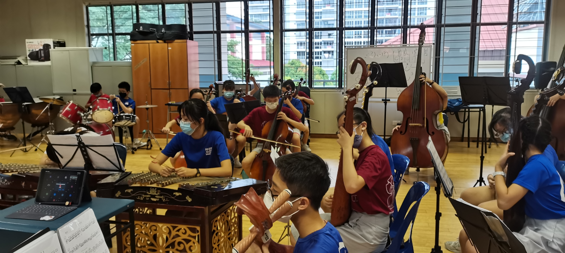 Students in blue and red shirts wearing masks rehearse with cellos, a guzheng, and other instruments in a music room.