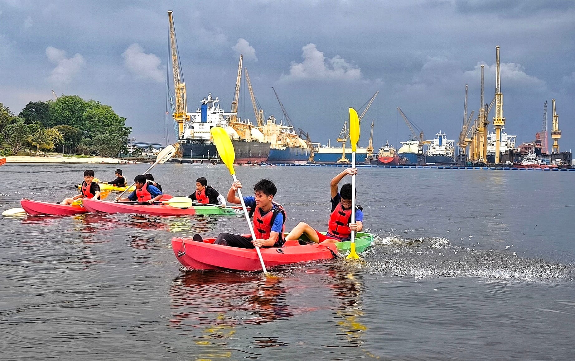 Several youths in red life jackets paddle kayaks on a harbor with large ships and cranes in the background.