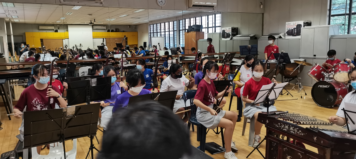 Students wearing masks rehearse traditional Chinese instruments including guzheng and drums in a music classroom.