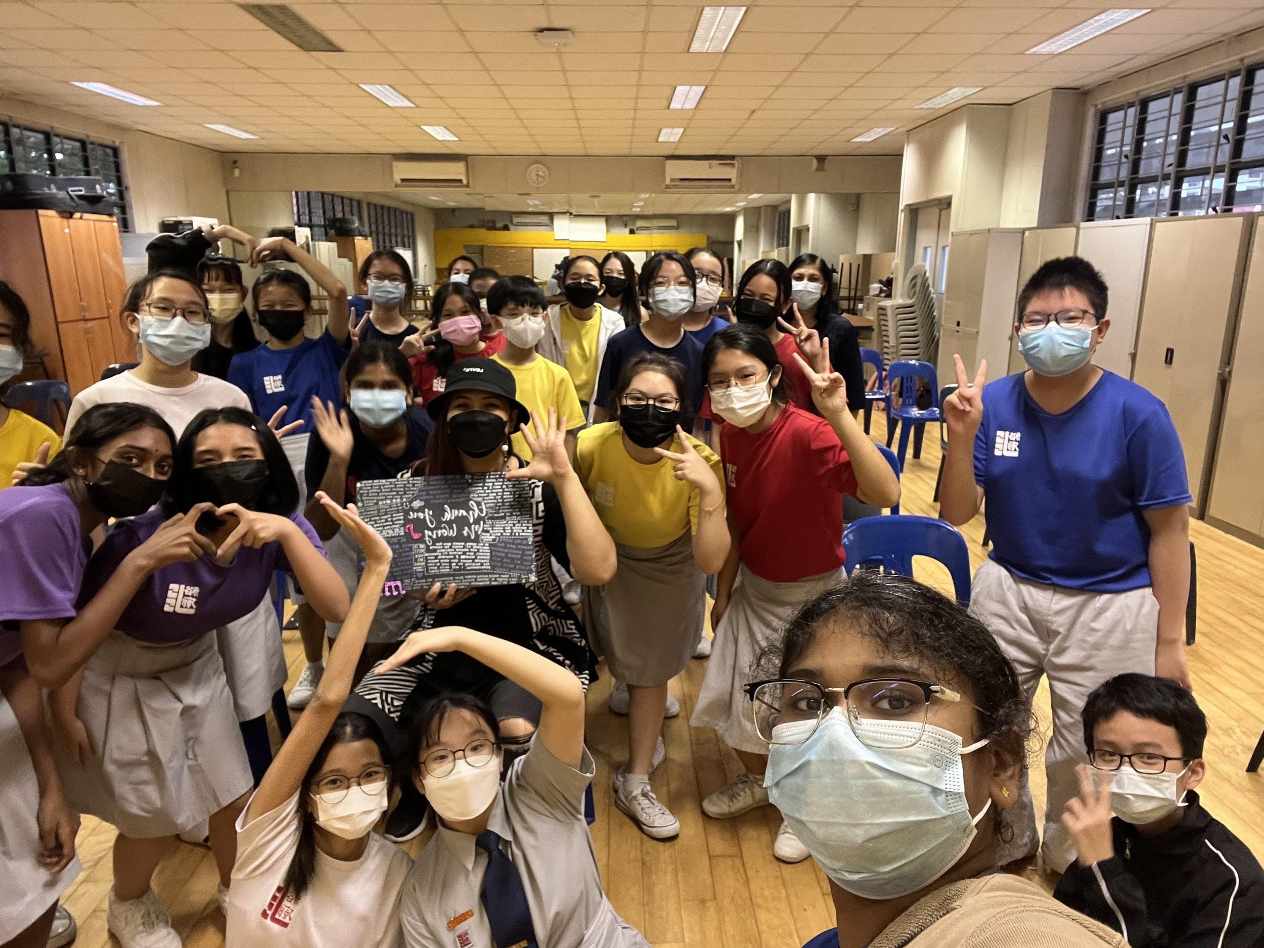 A large group of masked students pose together in a school hall, making peace signs and heart gestures.