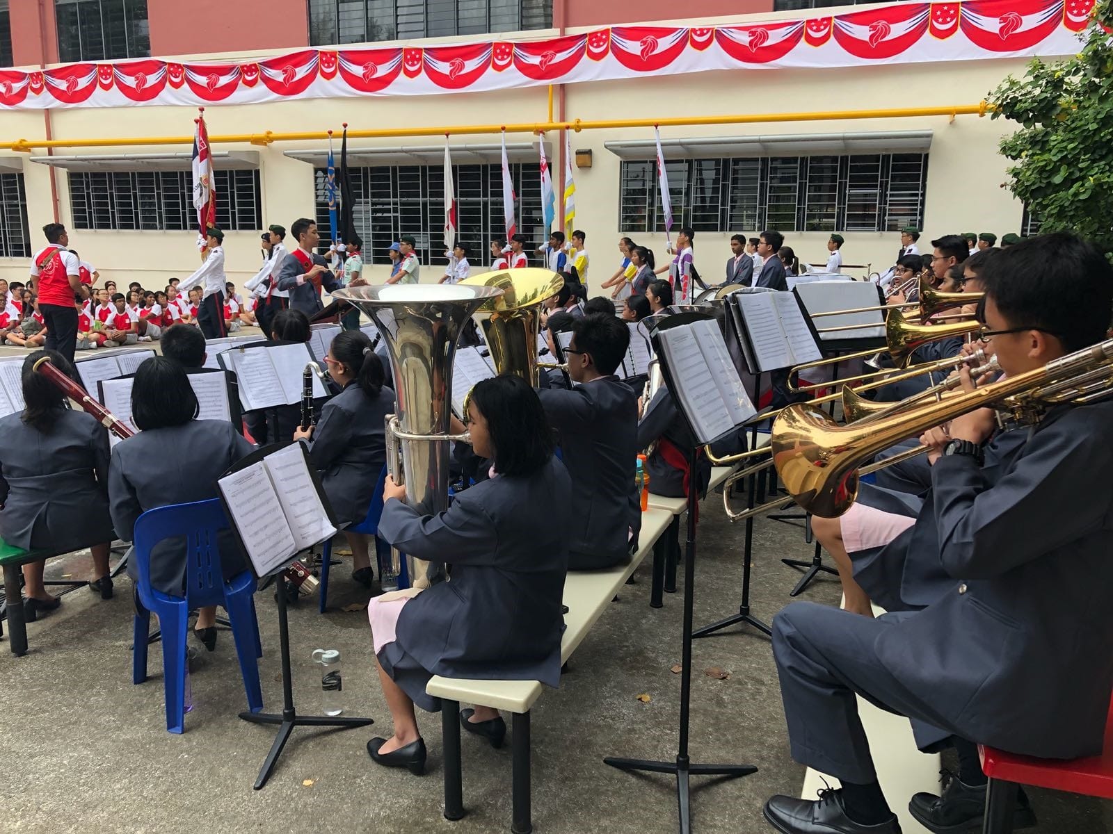School band in uniforms plays brass and wind instruments outdoors at a ceremony with red decorative banners above.