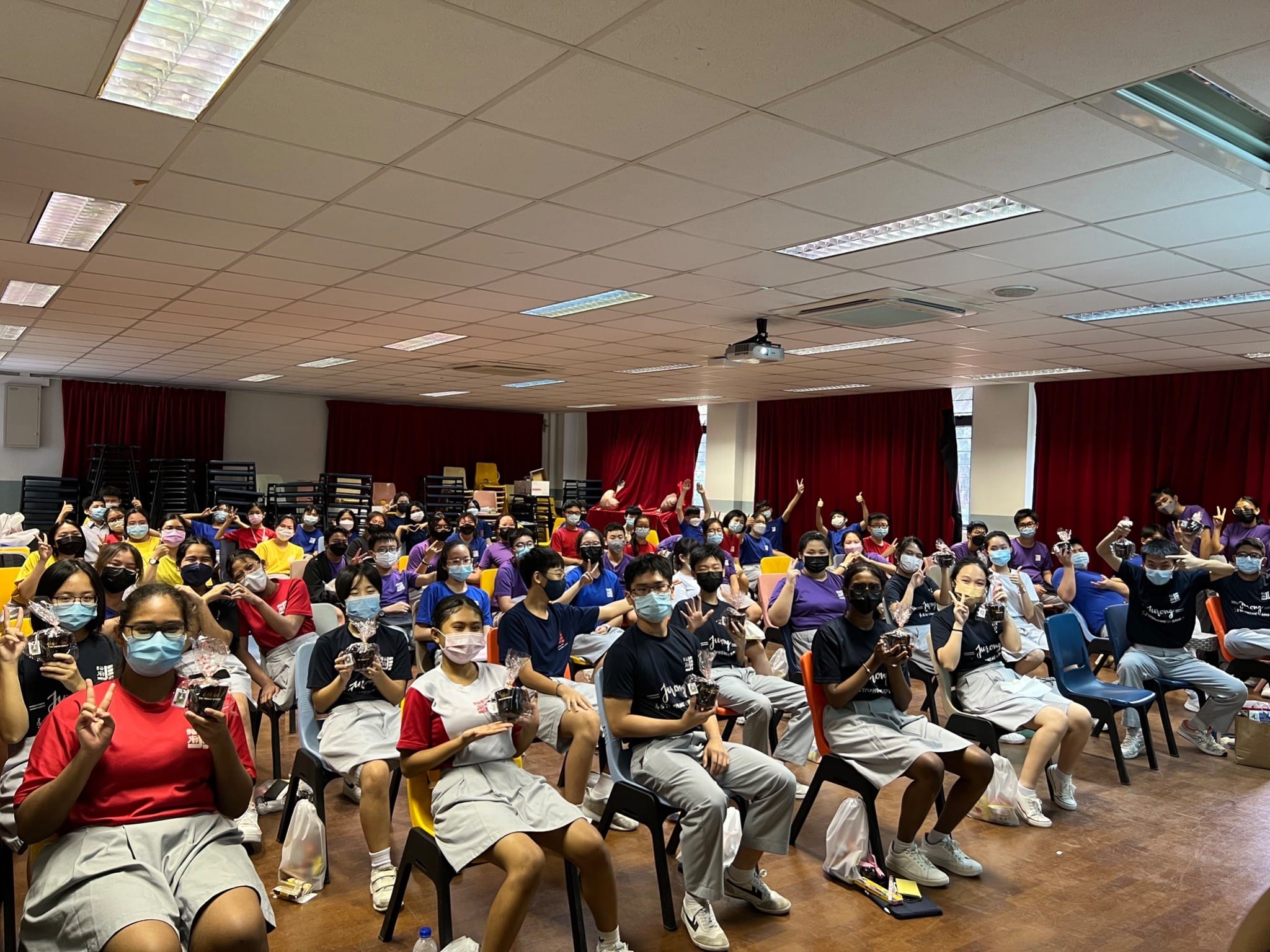 Large group of masked students seated in a hall, holding small items and posing with hand gestures.