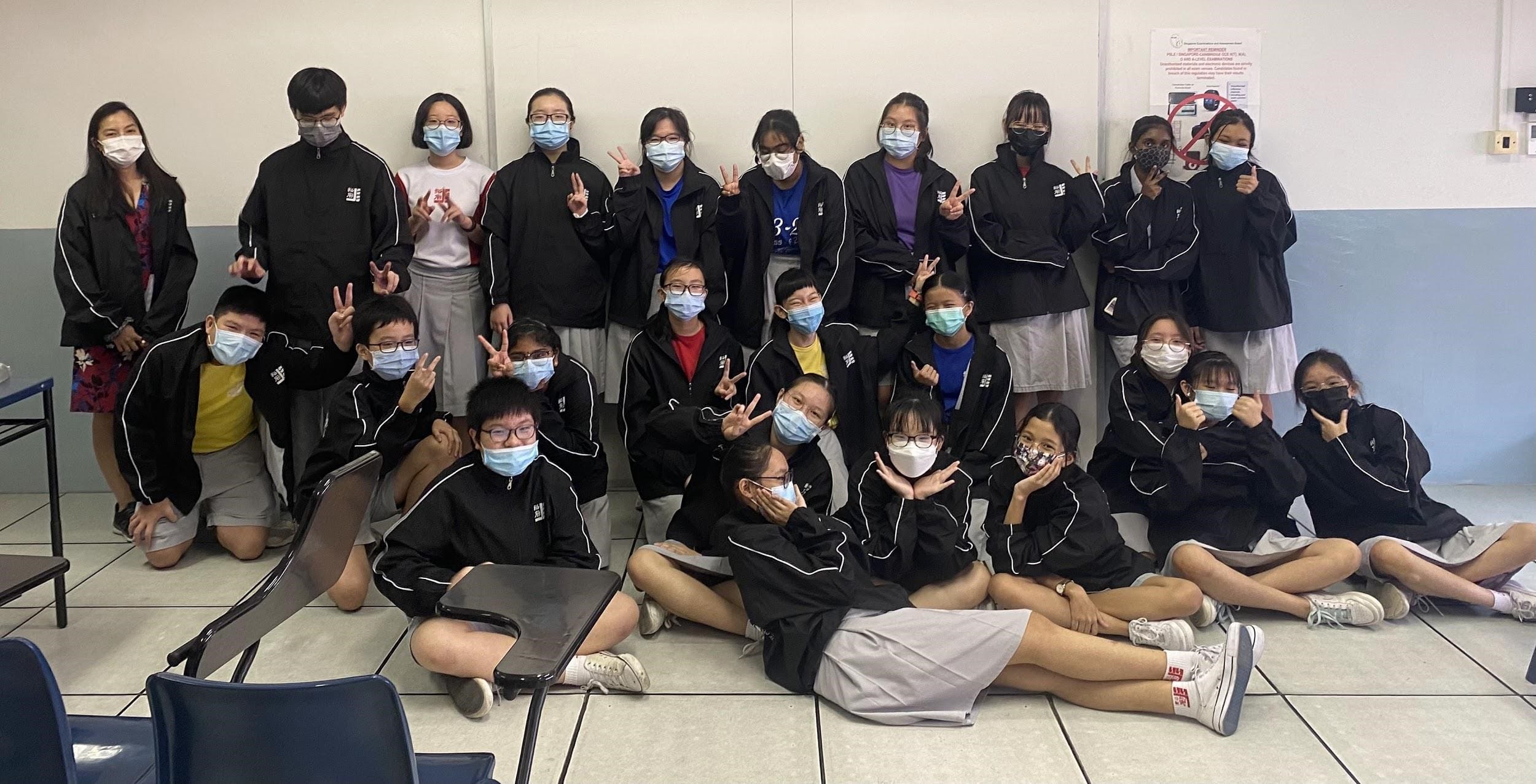A large group of students wearing black uniforms and face masks pose together in a classroom, making peace signs.