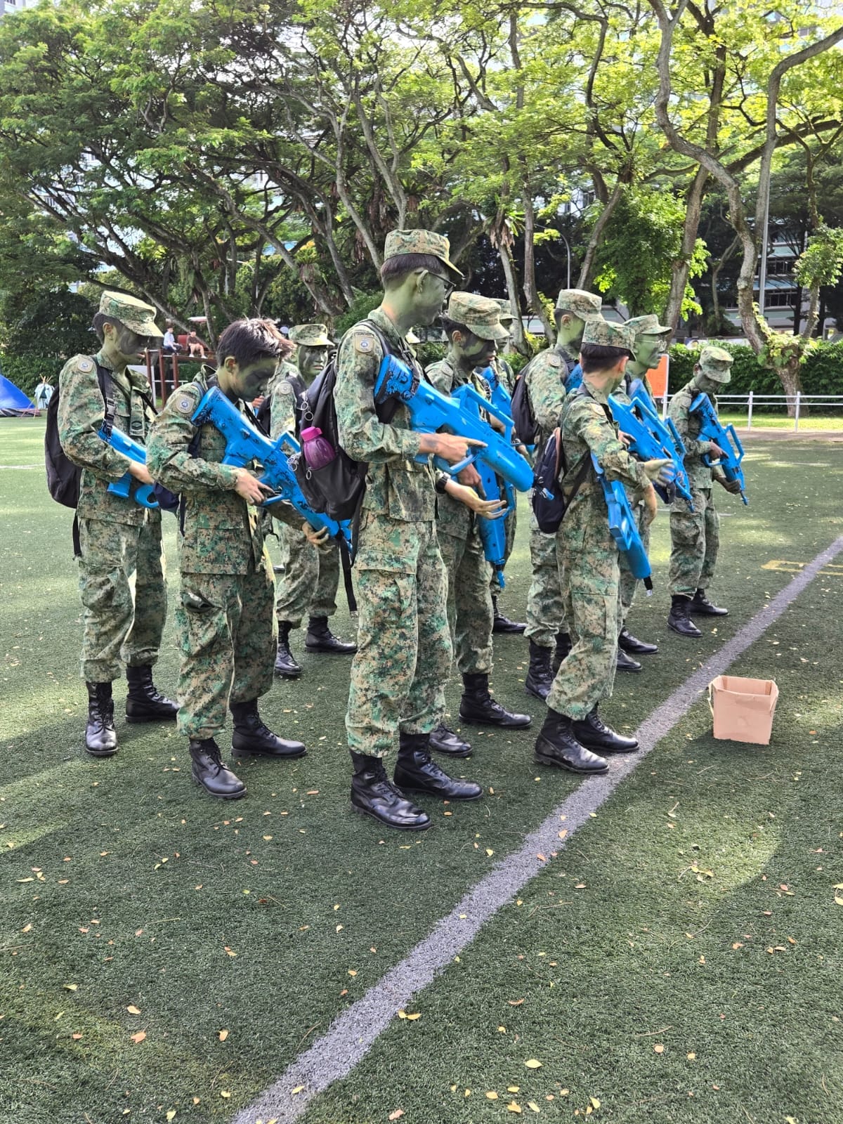 Soldiers in camouflage uniforms with green face paint hold blue training rifles on a grass field near trees.