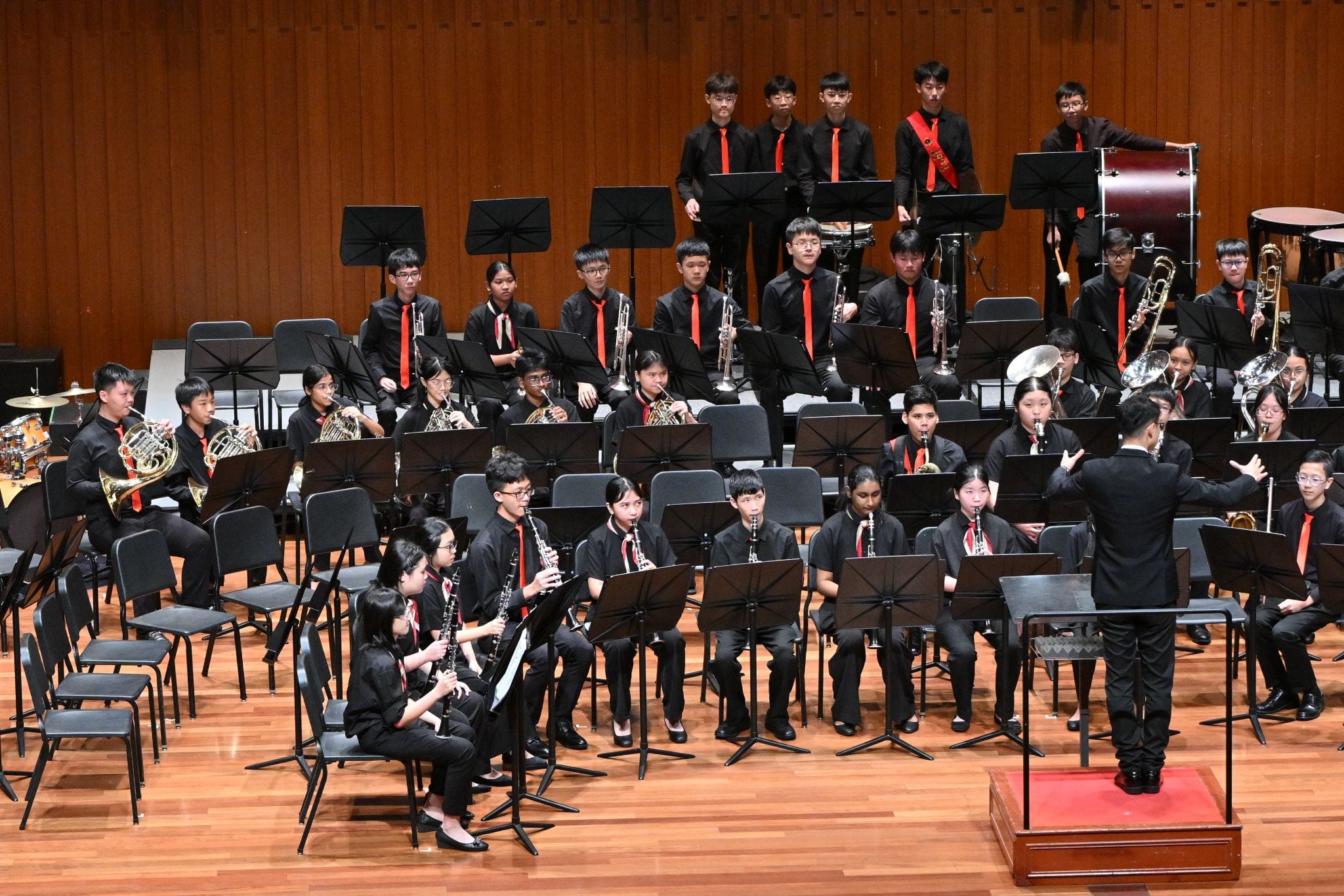 A youth wind band in black uniforms with red ties performs on stage, led by a conductor standing on a podium.