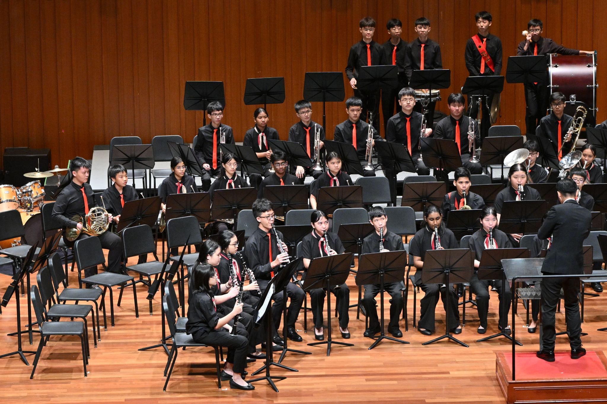 A youth wind band in black uniforms with red ties performs on a wooden stage, led by a conductor at the right.