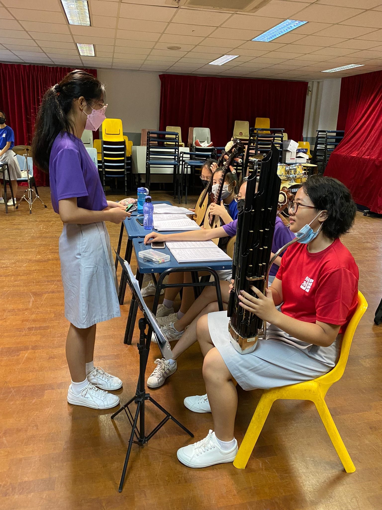 Students wearing masks practice traditional Chinese string instruments in a school rehearsal hall with red curtains.