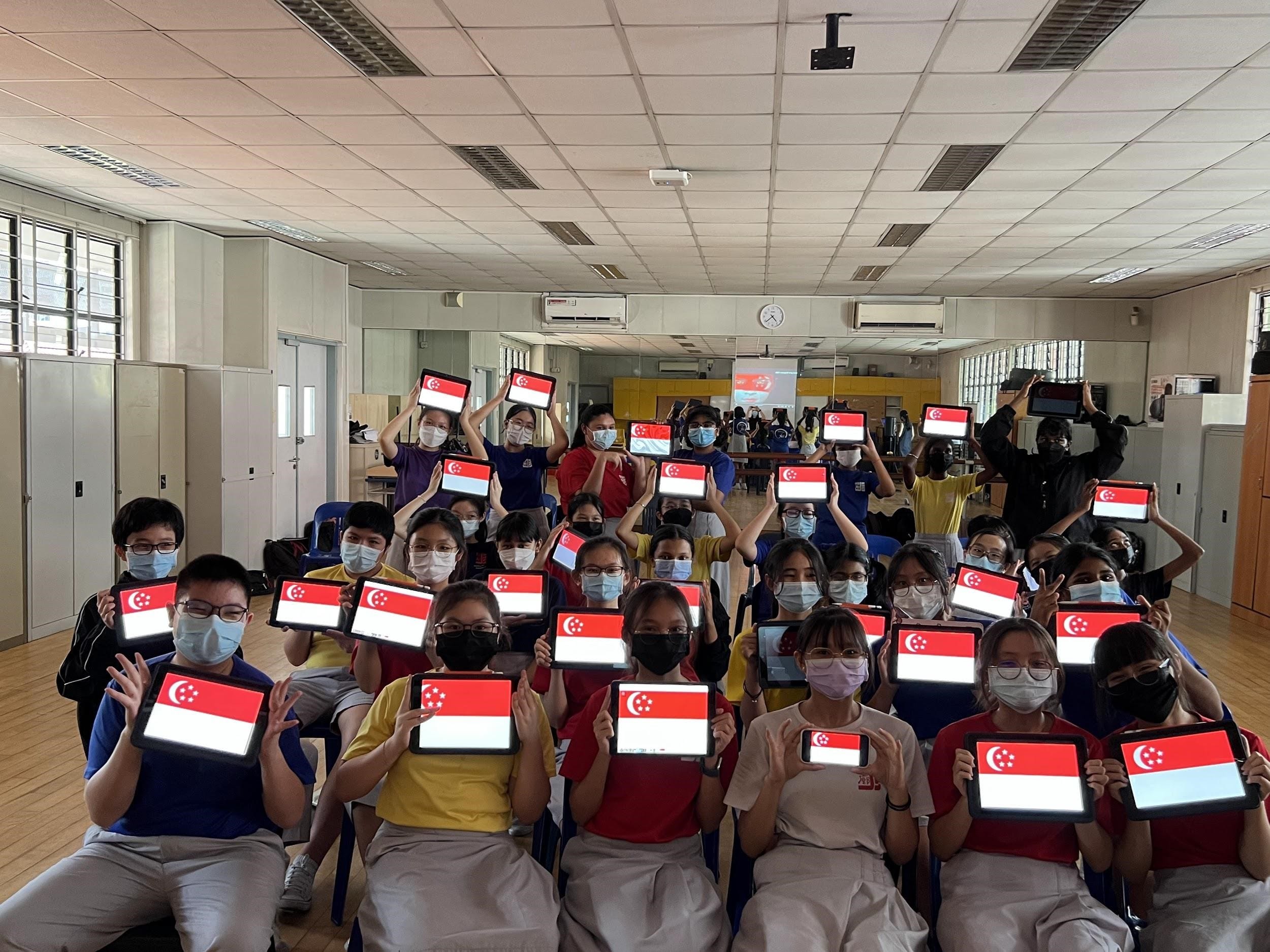 Students wearing masks hold up tablets displaying the Singapore flag in a classroom.