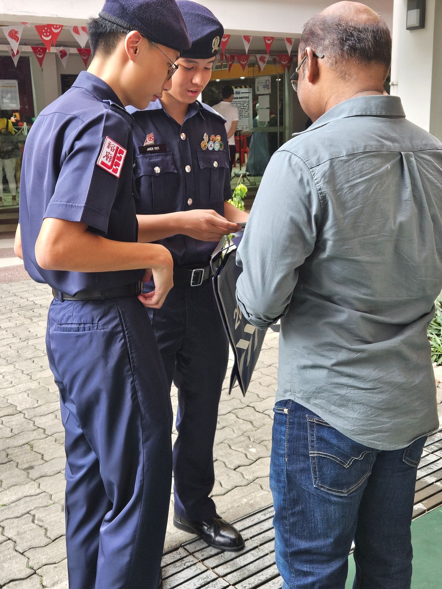 Two young officers in navy uniforms and berets interact with a man in a gray shirt outdoors on a paved area.