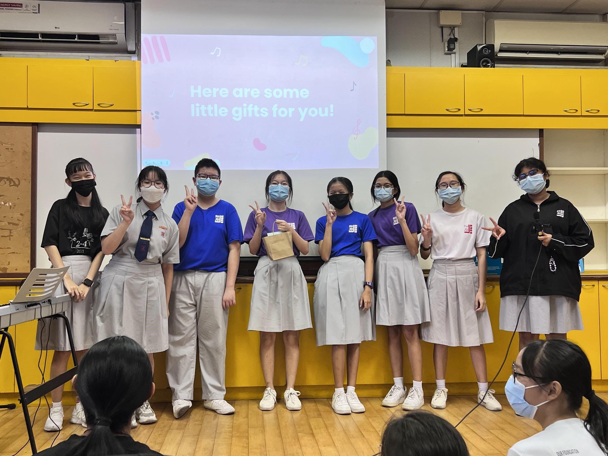 Eight masked students in school uniforms stand in a classroom making peace signs before a projected slide reading "Here are some little gifts for you!"