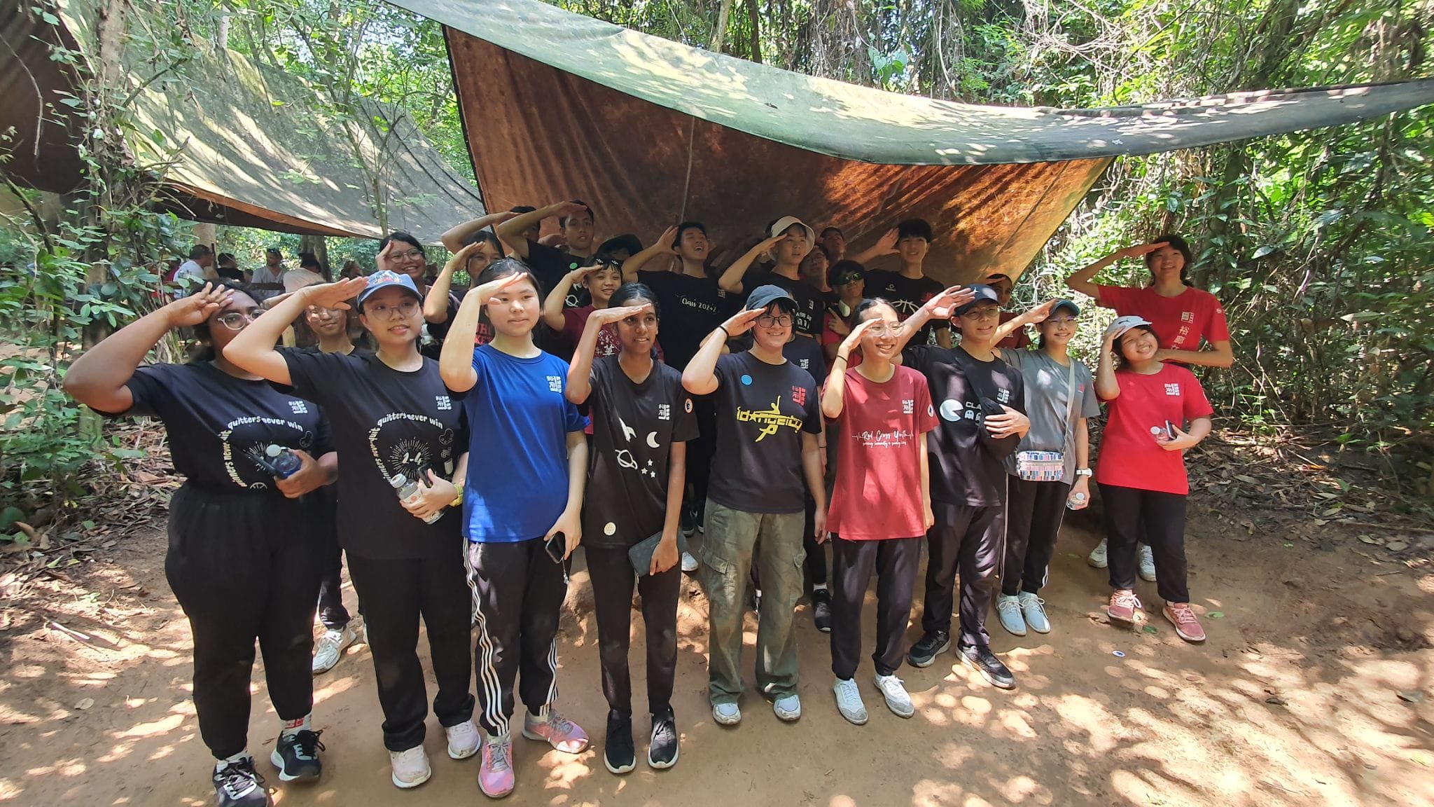 A large group of young people saluting together under a tarp shelter in a forested outdoor setting.