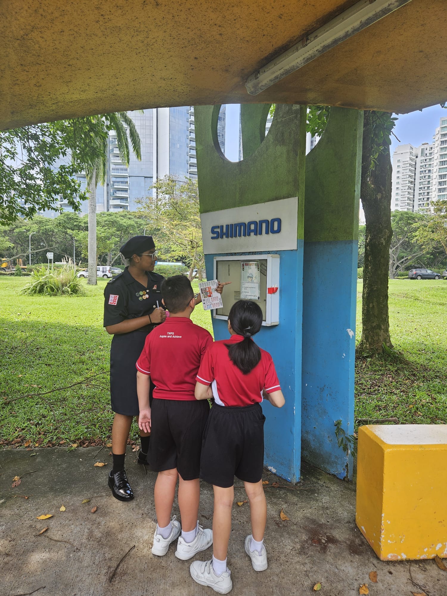An adult in black uniform guides two children in red shirts at a Shimano kiosk in a park.